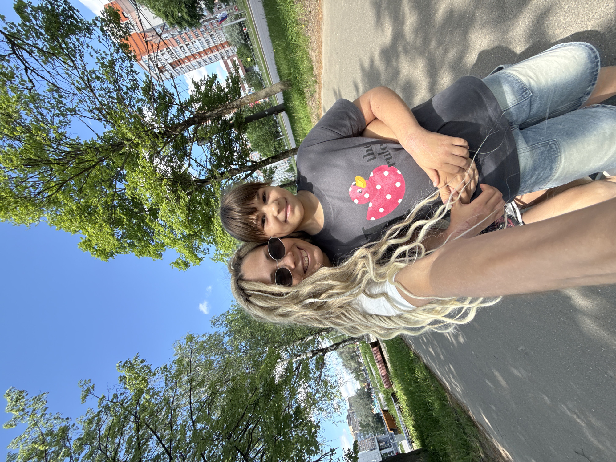The image depicts a woman with long, blonde, wavy hair and sunglasses taking a selfie with a child in an outdoor setting. The child is wearing a grey t-shirt with a red and white polka-dotted duck design. The woman is wearing a white top. They appear to be embracing. The background includes trees, grass, a paved pathway, and a multi-story building in the distance, aligning with a location in the city of Шебунино, Russia. The sky is blue with some clouds. The sun is shining brightly.
