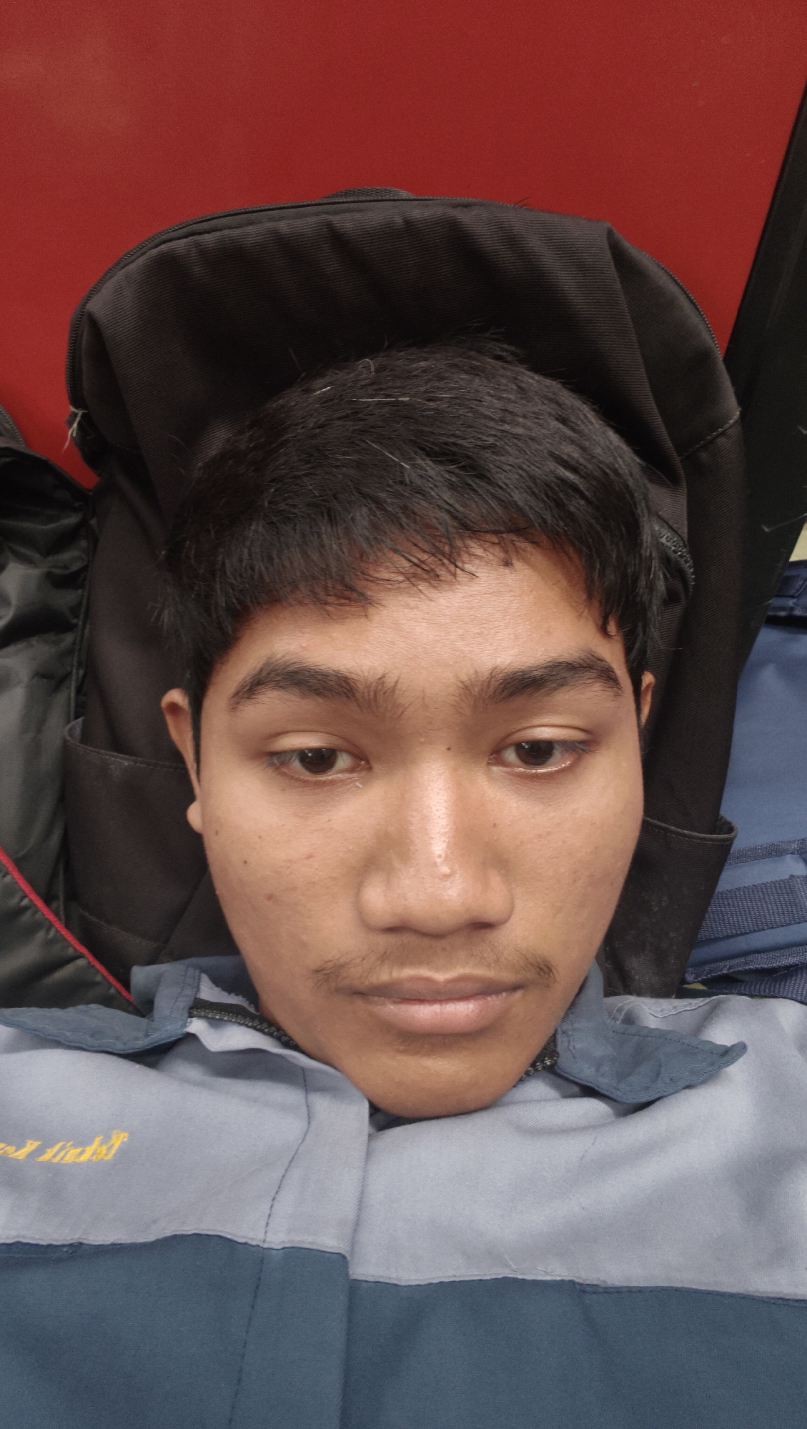 A young man is shown in a close-up, portrait-style photograph. He has dark hair, dark eyes, and appears to have a light mustache. He is wearing a gray and blue collared shirt. He is leaning against a black backpack, with a red background visible behind his head. The image was taken in Katrungan, Indonesia.
