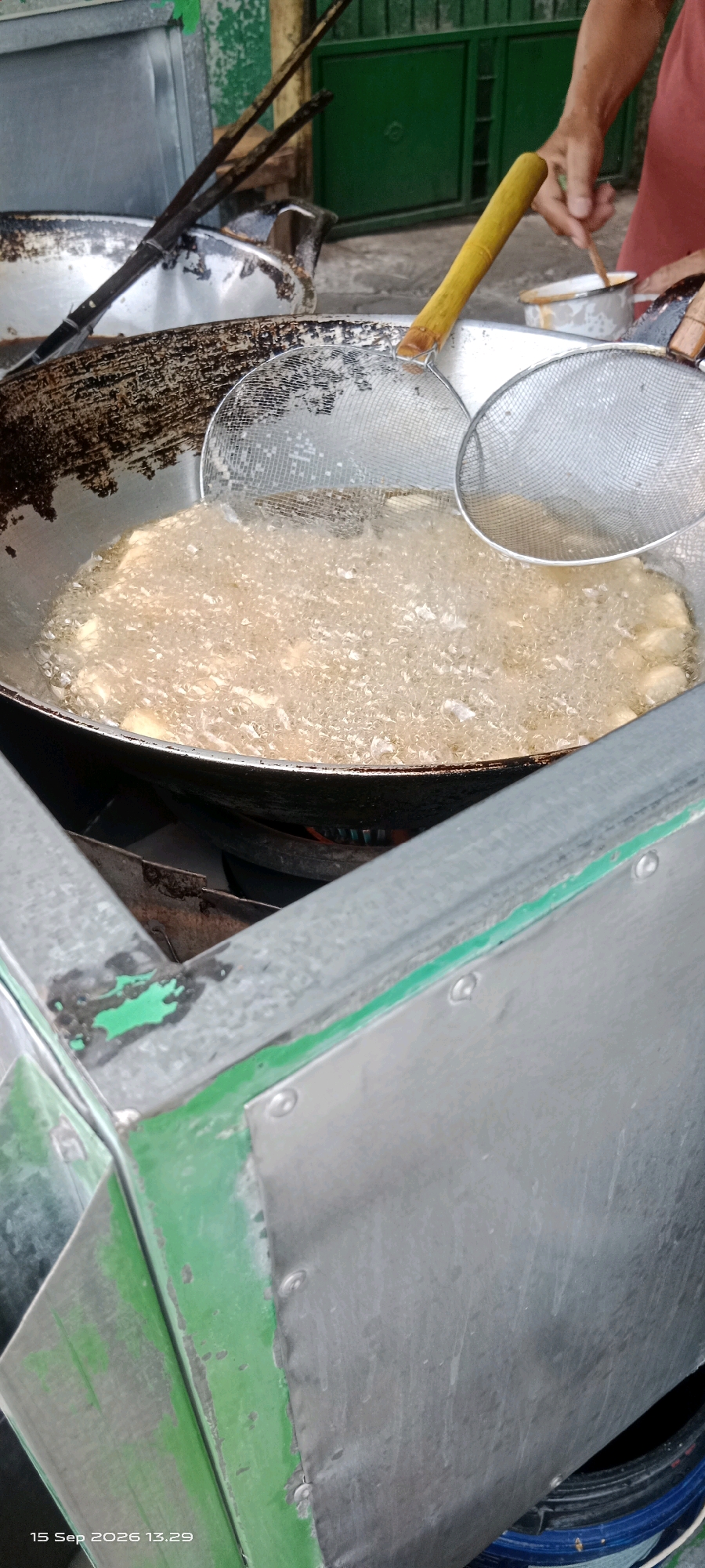 The image shows a close-up view of a street food vendor preparing food in what appears to be a public area in Kartasura, Indonesia. The primary focus is on a large, well-used wok filled with bubbling hot oil, in which pieces of food are being deep-fried. Two mesh strainers are being used to handle the food, one submerged in the oil and another held above it. In the background, a person's arms are visible, wearing a pink shirt, suggesting they are the vendor. They are holding a wooden utensil and a small metal bowl. The scene is set against a backdrop of a green metal gate and part of a food cart made of what looks like galvanized steel, showing signs of wear and tear. The lighting suggests it is daytime, possibly late morning or early afternoon, given the timestamp "15 Sep 2026 13:29" visible in the lower left corner. The overall impression is one of an active, bustling street food preparation environment.