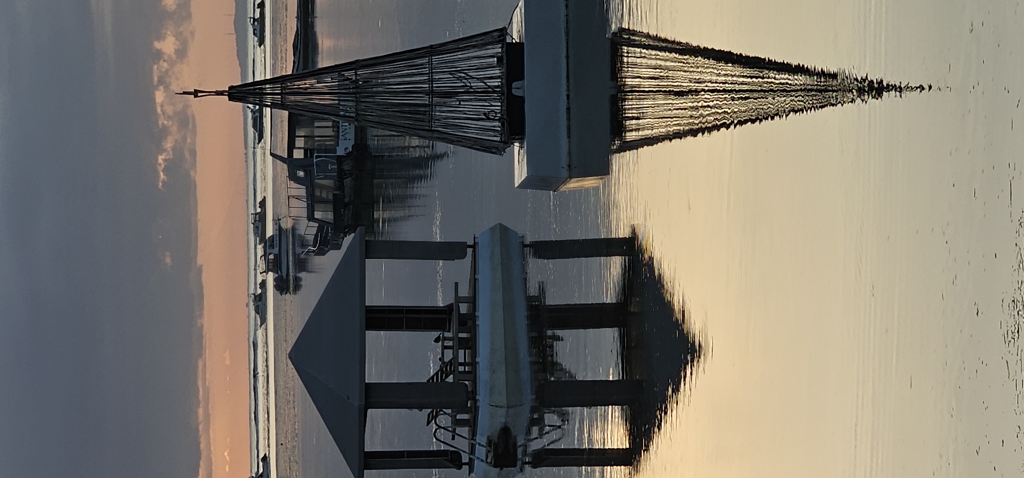 The image depicts a waterfront scene in Denpasar, Indonesia, captured during the early morning hours. The sun is rising on the horizon, reflected in the calm, slightly rippled water. Two prominent structures stand near the water's edge: a pier and a tower-like structure that may be part of a fishing net drying setup. Several boats are docked behind the pier. The sky is partly cloudy, with colors of the early morning light. The reflections of the structures and the light patterns in the water contribute to the overall composition of the scene.