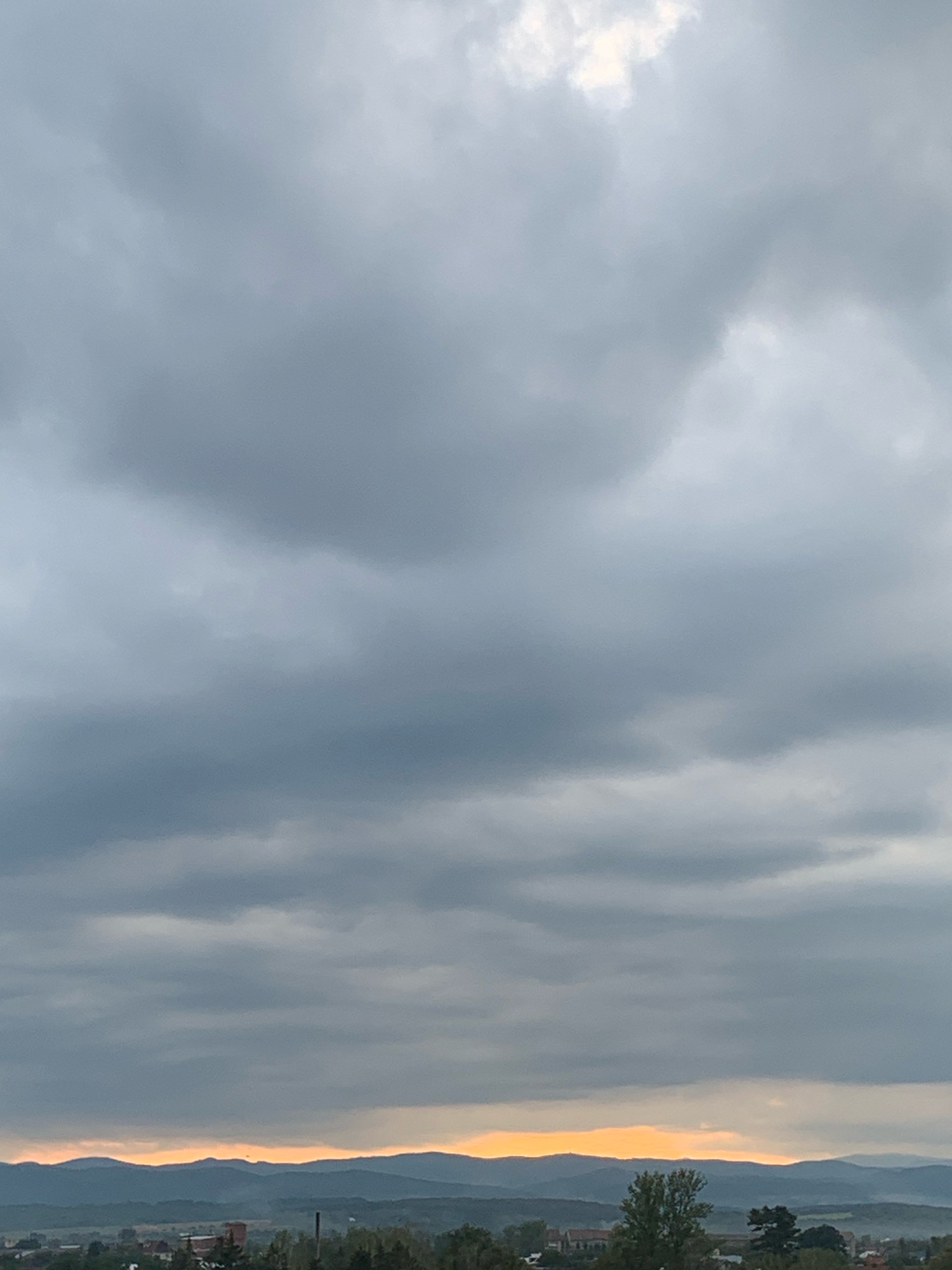 An overcast sky looms over a distant mountain range. Below the mountains, a small town is nestled, with trees and buildings dotting the landscape. The sky is a mix of grays and whites, with a sliver of orange light peeking through the clouds at the horizon. The overall scene is one of quiet contemplation, as the day transitions towards evening.