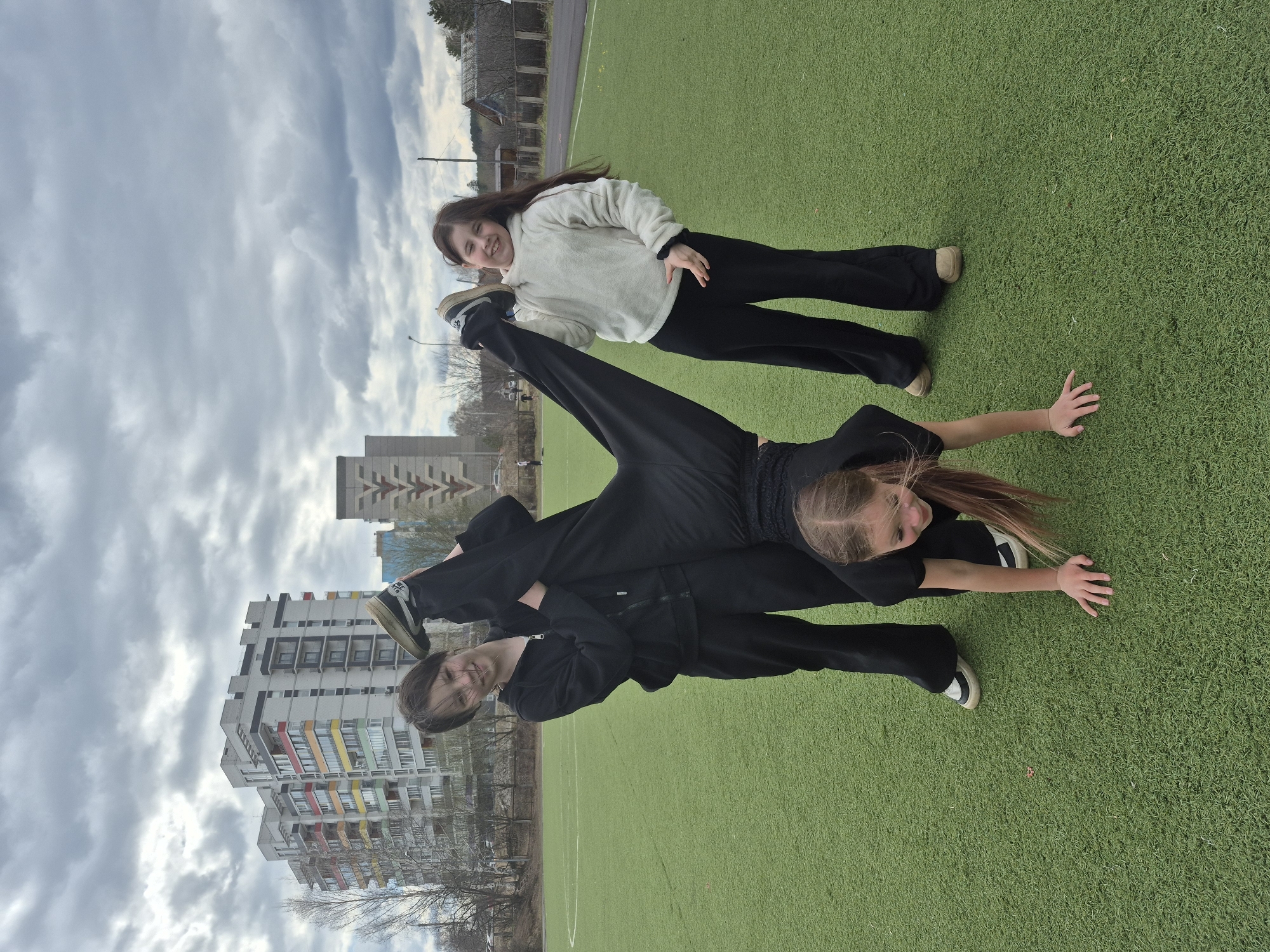 The image captures two young females on an artificial green turf sports field in Chaikovsky, Russia, under an overcast sky. The photograph is taken from a slightly elevated perspective, looking down and across the field.
One girl, positioned centrally, is performing a flexible, acrobatic pose. She is dressed in black athletic wear, including a long-sleeved top and pants. Her right leg is extended vertically towards the sky, while her upper body is close to the ground, supported by her hands. Her head is turned towards the camera, and she is smiling. Her long brown hair is visible, flowing slightly. On the side of her extended leg's shoe, the "NIKE" logo is visible.
Another girl stands behind the first, also smiling broadly and looking towards the camera. She is wearing a light-colored (cream or off-white) fuzzy sweater and black pants, with dark sneakers. She stands upright, with one hand casually placed on her hip.
The background features several large, multi-story apartment buildings, some with colorful accents on their balconies, characteristic of Soviet-era architecture. Bare trees suggest either late autumn, winter, or early spring. A fence runs along the edge of the field, and a few utility poles are also visible. The sky is filled with heavy, grey clouds, indicating an overcast day with diffused light. The overall mood appears joyful and active.