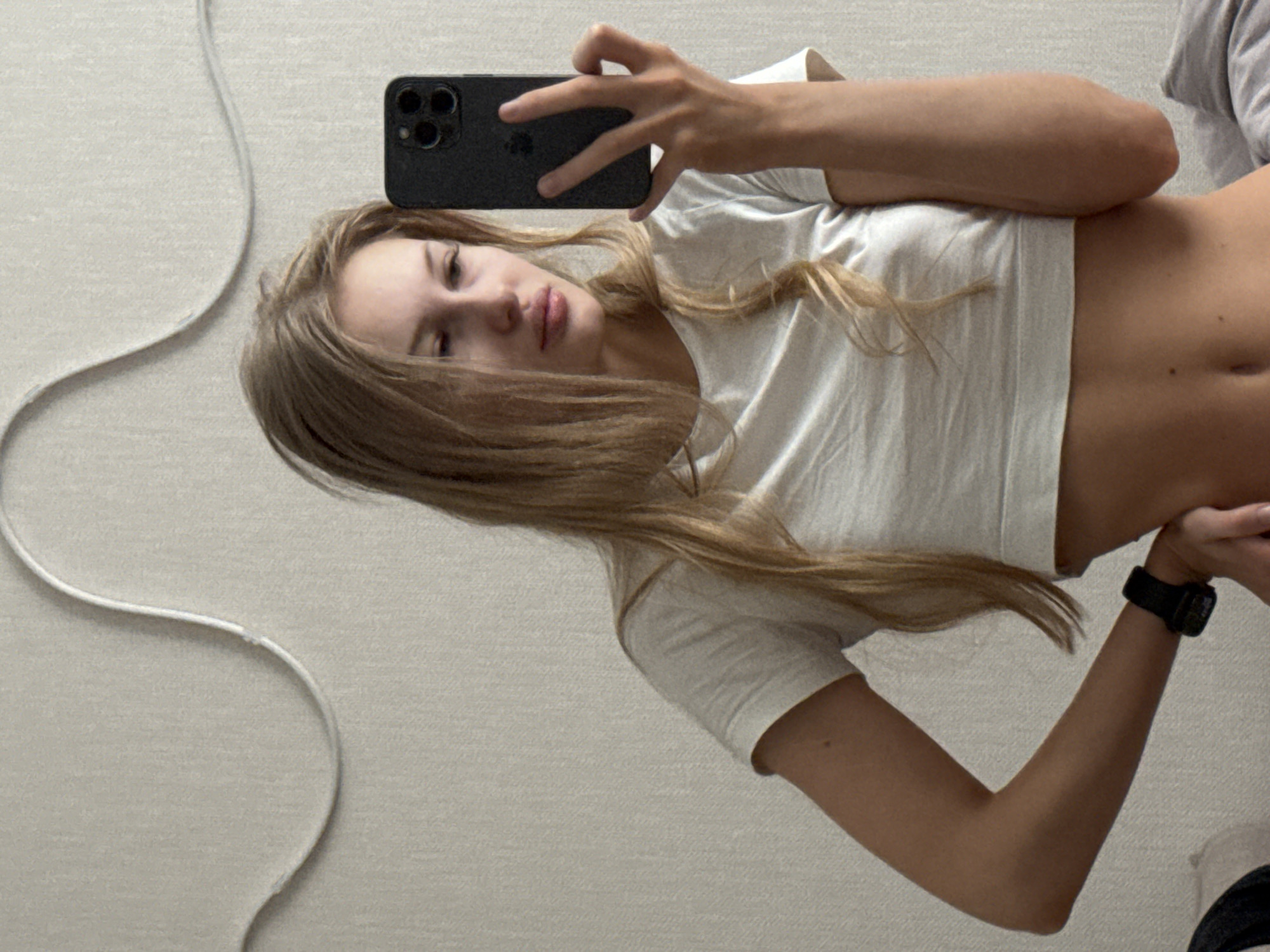 A young woman with long blonde hair takes a selfie in a room. She is wearing a white t-shirt that exposes her midriff and a black watch on her left wrist. The background is a textured wall with a curved white decorative element. The image is taken indoors, likely in a residential setting, and there is no indication of the city of Perm, Russia, or any specific urban location.