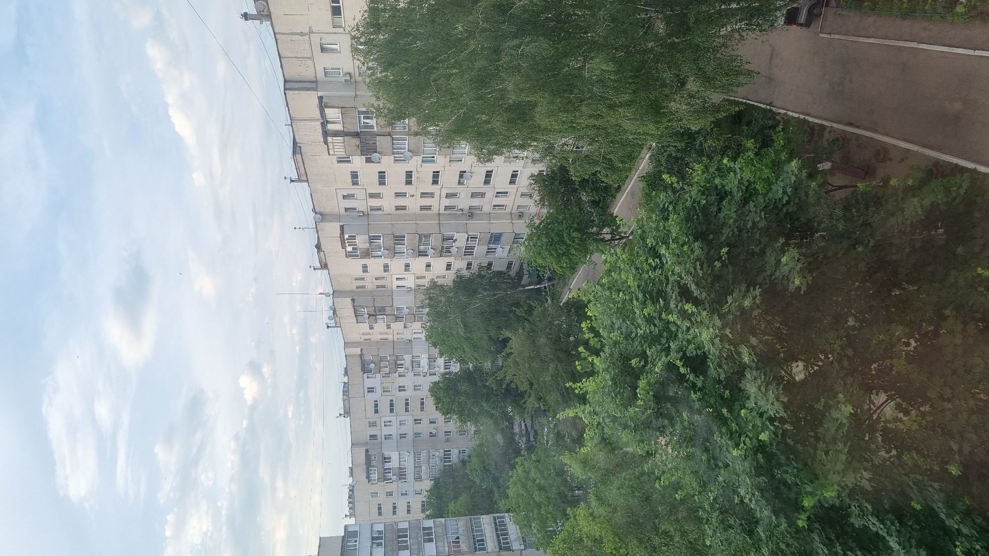 The image depicts a view of an urban environment in Smila, Ukraine, characterized by residential apartment buildings and lush greenery. The buildings are multi-story, with light-colored facades and numerous windows, suggesting a densely populated area. The architecture appears typical of Soviet-era housing blocks, with balconies and external air conditioning units visible. Surrounding the buildings are numerous trees, some tall and mature, others smaller and more bushy, creating a strong contrast between the man-made structures and the natural elements. The sky is partly cloudy, with soft white clouds against a pale blue backdrop, indicating fair weather. A paved walkway or road runs alongside the buildings, bordered by a strip of grass and trees. A dark-colored vehicle is parked along the roadside. The overall impression is one of a typical residential neighborhood within a Ukrainian city, balancing urban development with abundant vegetation.