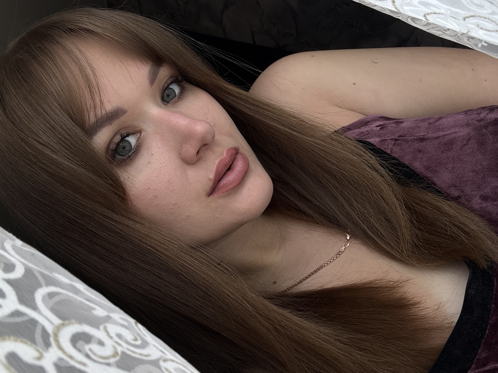 A woman is lying on a bed, resting her head on a pillow. She has light brown hair with bangs, light skin, and light-colored eyes. She is wearing a maroon top with black trim. She is also wearing a gold necklace. The pillow has a gray and white pattern. The background is dark, with some light coming from above. The location is unknown.