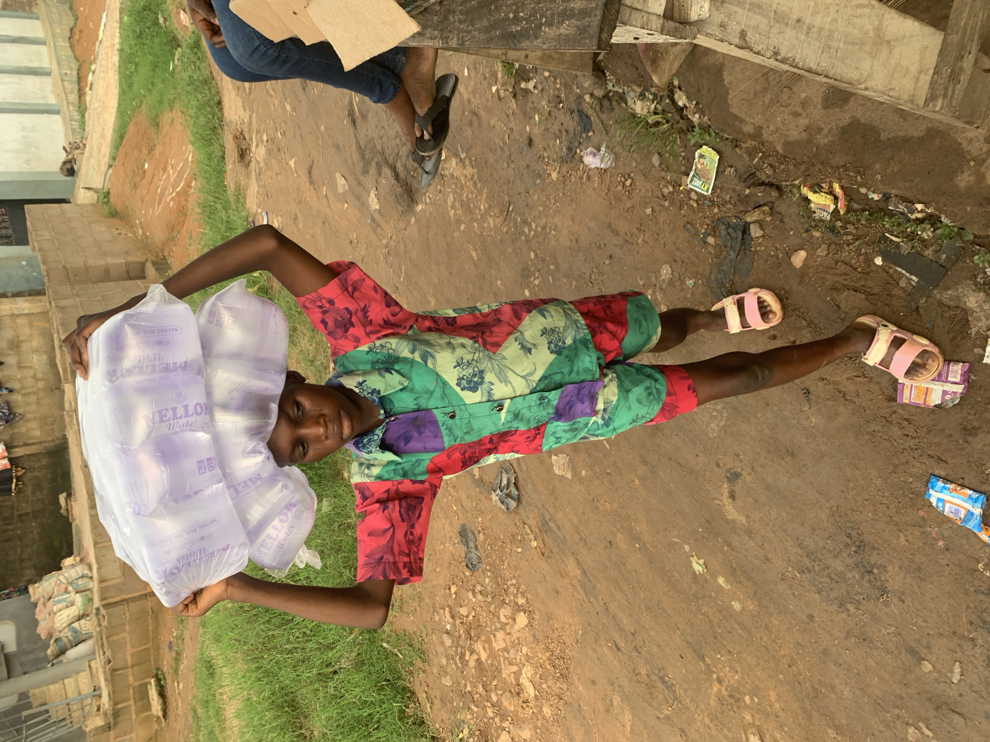 A young person with dark skin and a colorful patterned shirt and shorts carries a large stack of bagged water on their head while walking on a dirt path. The person is wearing pink sandals. In the background, there are buildings, grass, and another person sitting down wearing blue jeans and flip-flops. Trash is scattered on the ground. This scene is located in the city of Shagamu, Nigeria.