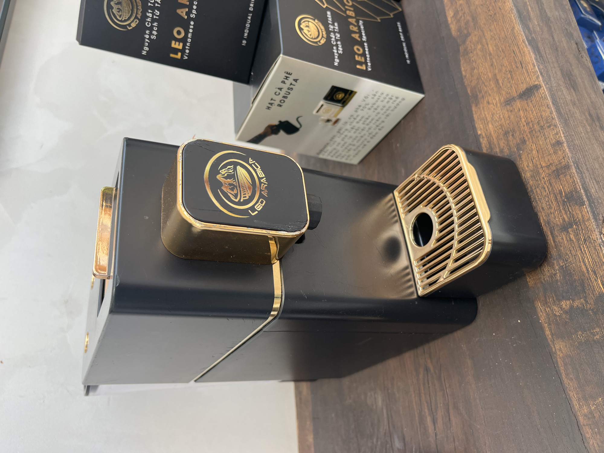The image shows a black and gold coffee machine, with a square hopper on the left and a slatted drip tray on the right. Two black and white boxes of LEO ARABICA Vietnamese coffee are behind the machine. The coffee machine is sitting on a dark wooden surface. The background is a white and gray marble-like surface. The location is assumed to be in Bac Nha Trang Ward, Vietnam, based on the coffee brand.