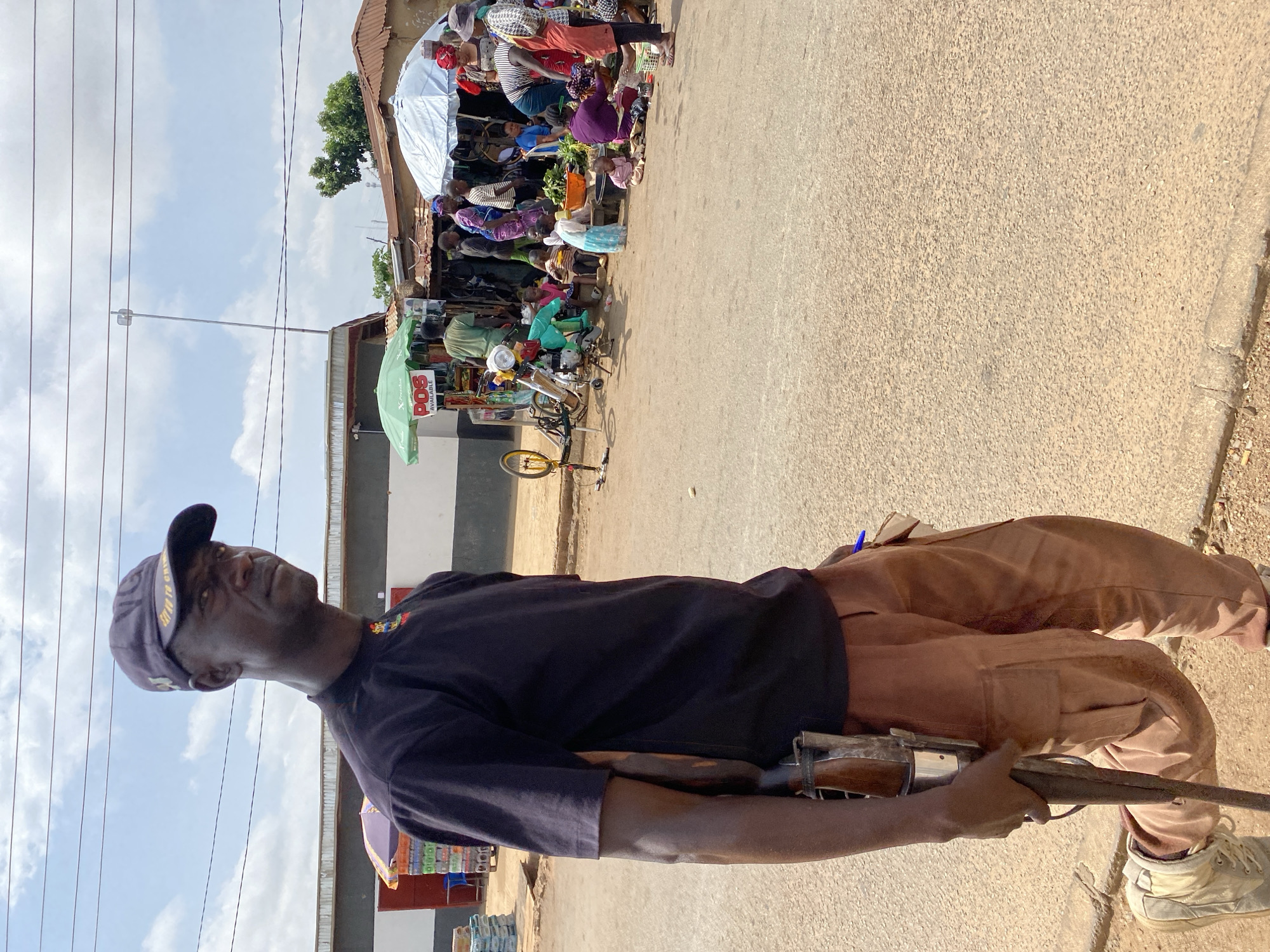 A man wearing a dark t-shirt, brown cargo pants, and a dark baseball cap stands on a paved road. He is holding a rifle with a wooden stock down his right side, his right hand gripping the fore-end and his left hand resting on the trigger guard. His left foot is slightly forward, and he appears to be walking or posing. In the background, a bustling outdoor market is visible. Stalls with umbrellas are set up, displaying various goods. Several people, including women and children, are engaged in activities such as shopping, selling, and sitting. Some children are playing on the ground. The environment suggests a sunny day in a Nigerian town.