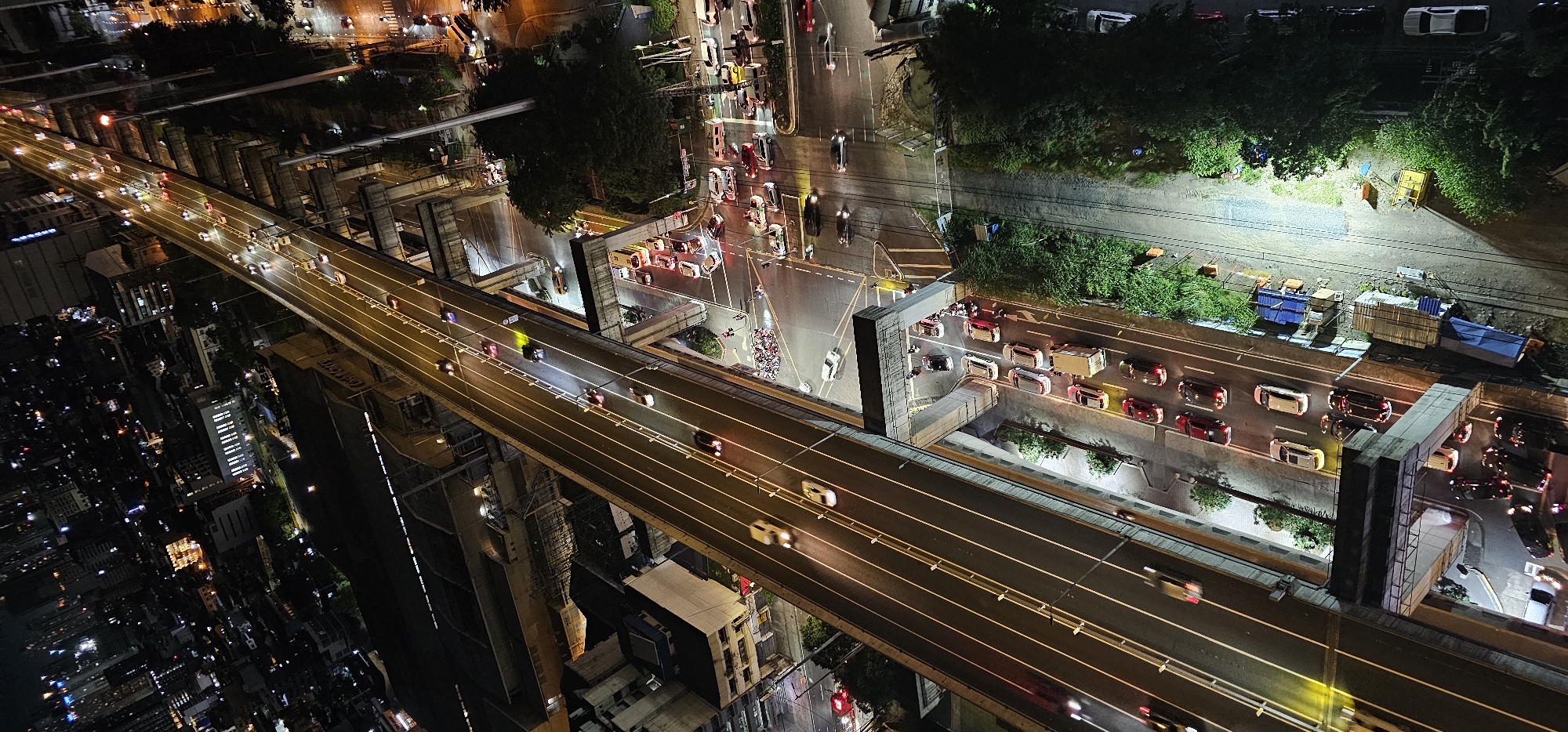 This is an aerial nighttime view of a highway in Makati, Philippines, with a city skyline visible in the background. The highway is elevated and illuminated by streetlights, with multiple lanes of traffic. Cars, buses, and motorcycles are visible on the highway and on the streets below. Buildings and trees line the sides of the highway, and construction materials are present in some areas. The scene appears to be a busy urban environment at night.