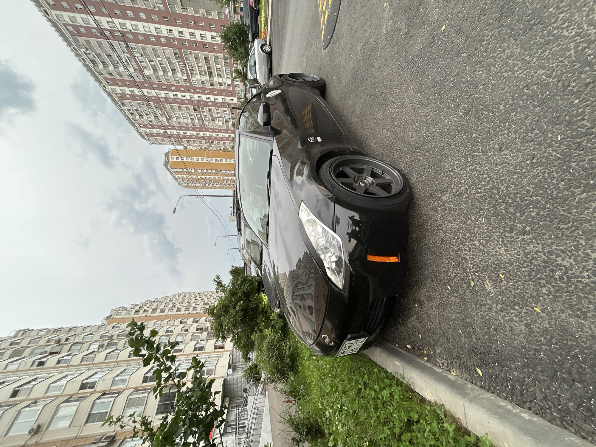 A black Nissan 350Z sports car is parked on an asphalt road next to a curb with grass and bushes. The car has custom gray rims. In the background, there are tall, multi-story apartment buildings typical of the Moskovskiy city, Russia. The sky is overcast with clouds. A street lamp is visible on the left side of the image.