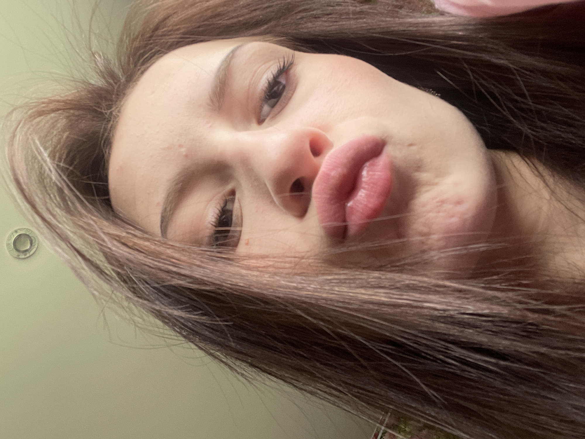 A close-up, low-angle selfie captures a young person, likely female, with long brown hair, posing indoors. The subject is looking down towards the camera, her face tilted, making a playful "pouty" expression with puckered lips. Her long, dark brown hair frames her face, with strands visible across the image. The background consists primarily of a plain, light green wall. In the upper left corner, a small, round metallic fixture, possibly a screw head or grommet, is visible on the wall. A hint of pink fabric is subtly visible in the upper right background. No text is visible.