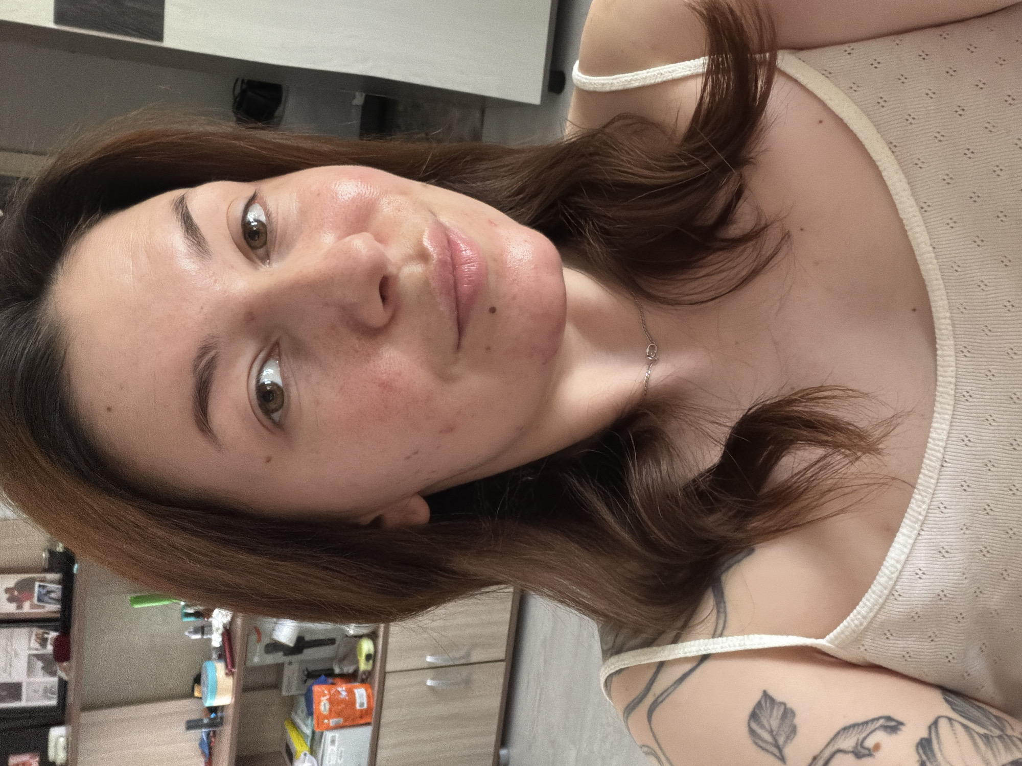 A selfie of a woman with brown hair and hazel eyes. She is wearing a white tank top and a silver necklace. She has fair skin with some redness and blemishes. The background appears to be the interior of a home, with a cabinet and miscellaneous items visible. There's no specific information available to confirm the exact location within the city of Chaikovsky, Russia.