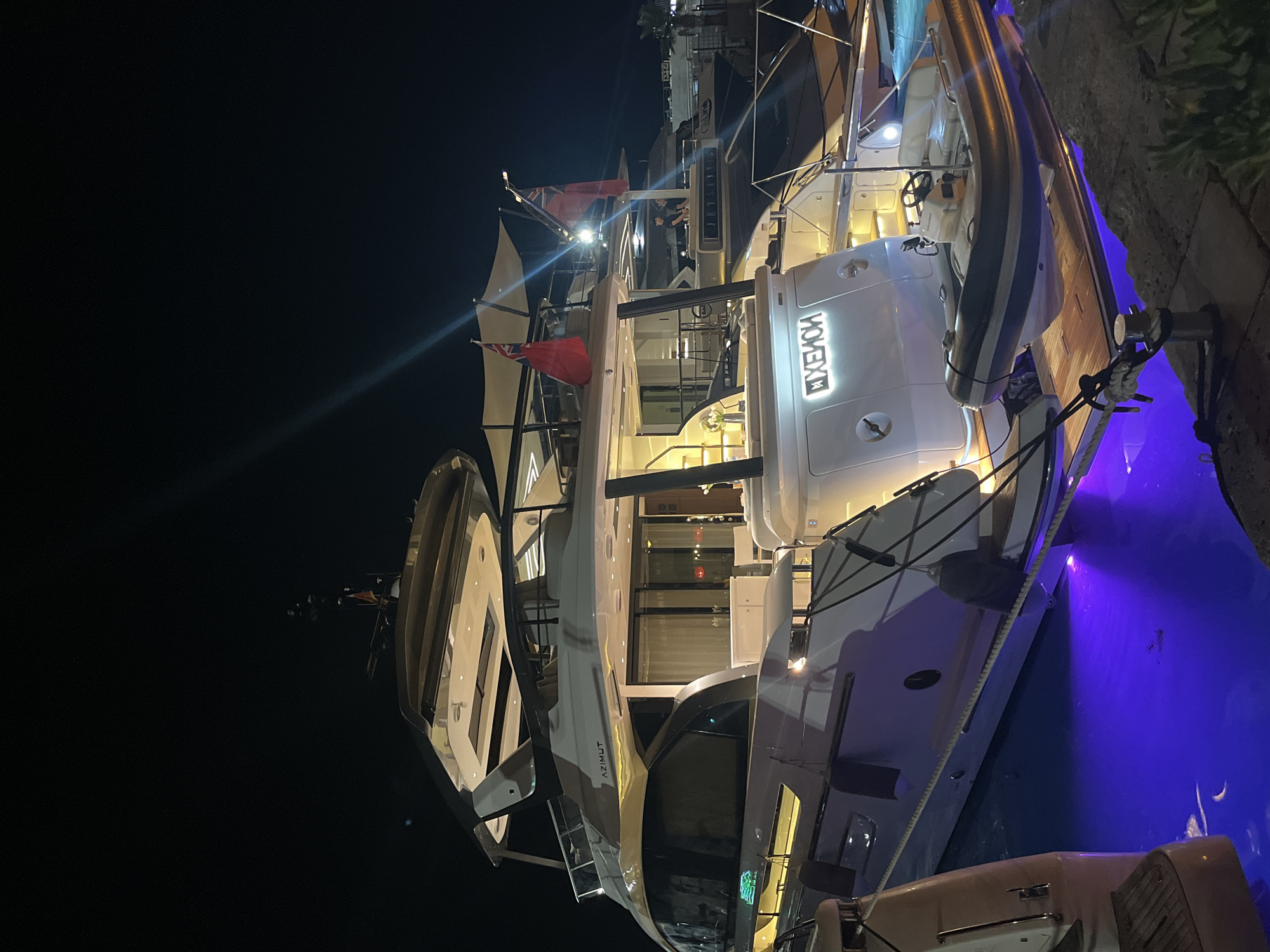 A luxury Azimut yacht, named "XENION," is docked at night in Budva, Montenegro. The yacht is illuminated by interior and exterior lights, with blue underwater lights reflecting on the water. A smaller dinghy is attached to the stern of the yacht. Two flags, one British, are visible on the upper deck. The scene is dark, with only the yacht and its immediate surroundings illuminated. There are no people clearly visible on the yacht, but two individuals are seen on the deck of another yacht in the background. The yacht is moored to a stone quay.