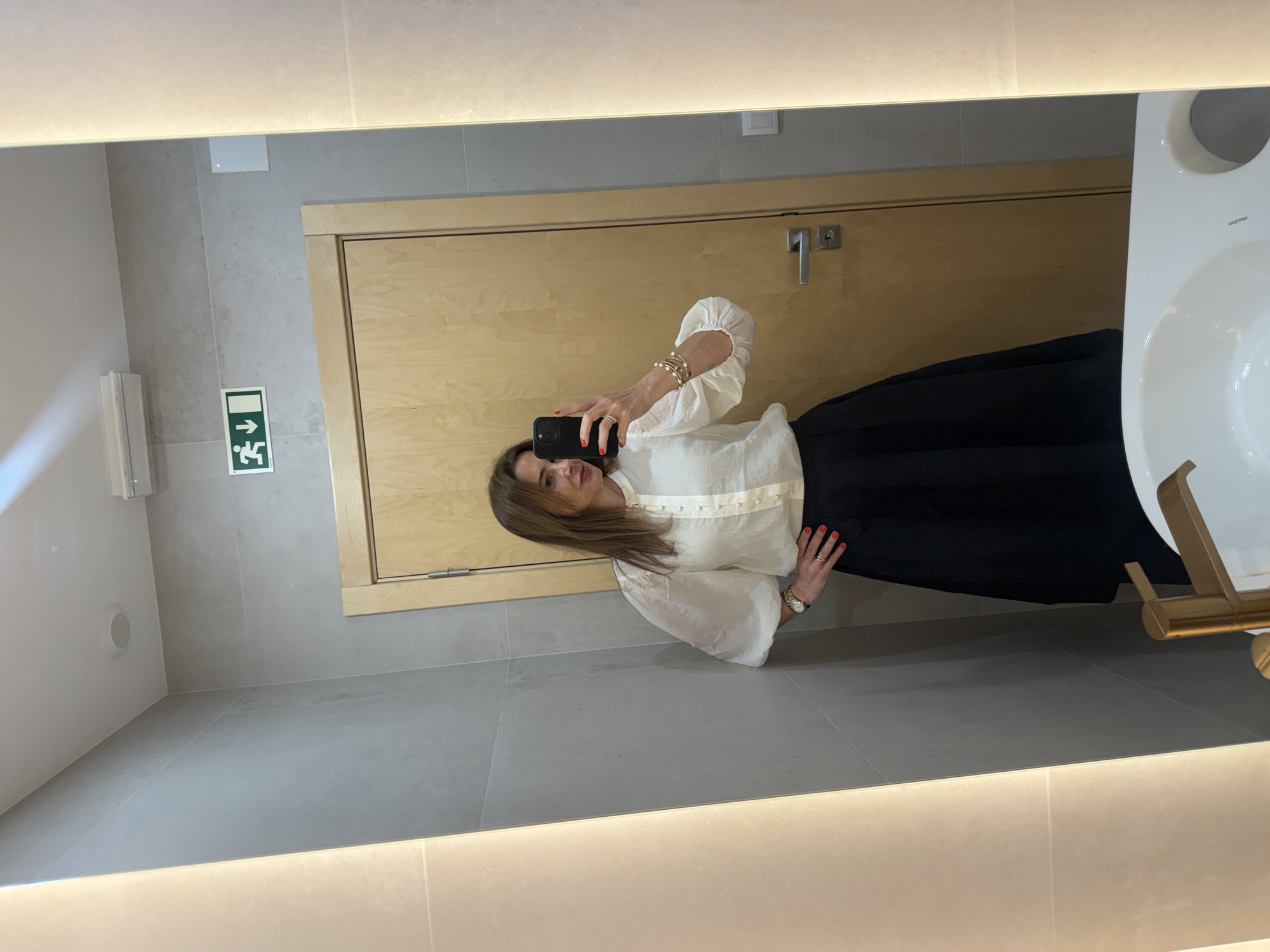 A woman is taking a selfie in a bathroom. The bathroom features gray tiles, a wooden door, and a white sink. An emergency exit sign is visible on the wall. The woman is wearing a white blouse and a black skirt. She is holding a black smartphone to capture the image. While the location is a bathroom in Torres Vedras, Portugal, there is no additional information to deduce from the image.