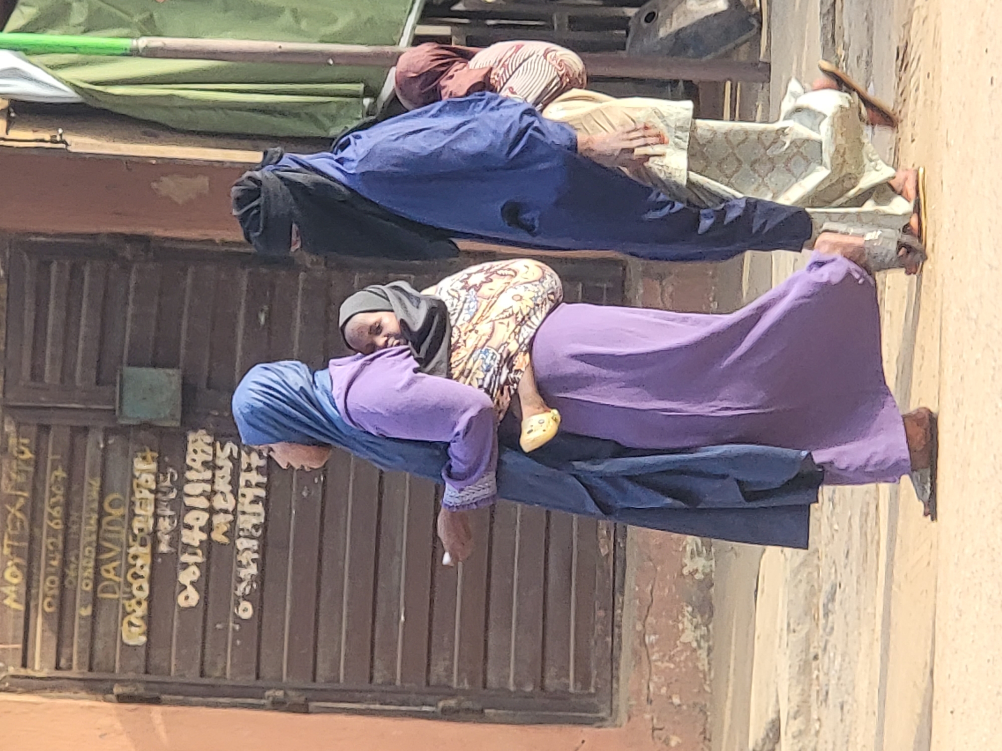 The image captures a street scene in Maiburiji, Nigeria. A woman in a purple dress and blue headscarf carries a child on her back, with the child wearing a patterned top, black head covering, and yellow shoes. The woman holds a white object in her hand. To the left, a metal gate or door bears faded yellow writing including the words "MOYTEX PETETAT" and the name "DAVIDO" along with phone numbers and other text. Further back, another woman is visible, wearing a blue garment and black head covering, with a third woman wearing a brown head covering and patterned shirt is lying next to her. Above them is a green awning. The scene appears to be on a sunny day with a building adjacent to the street.