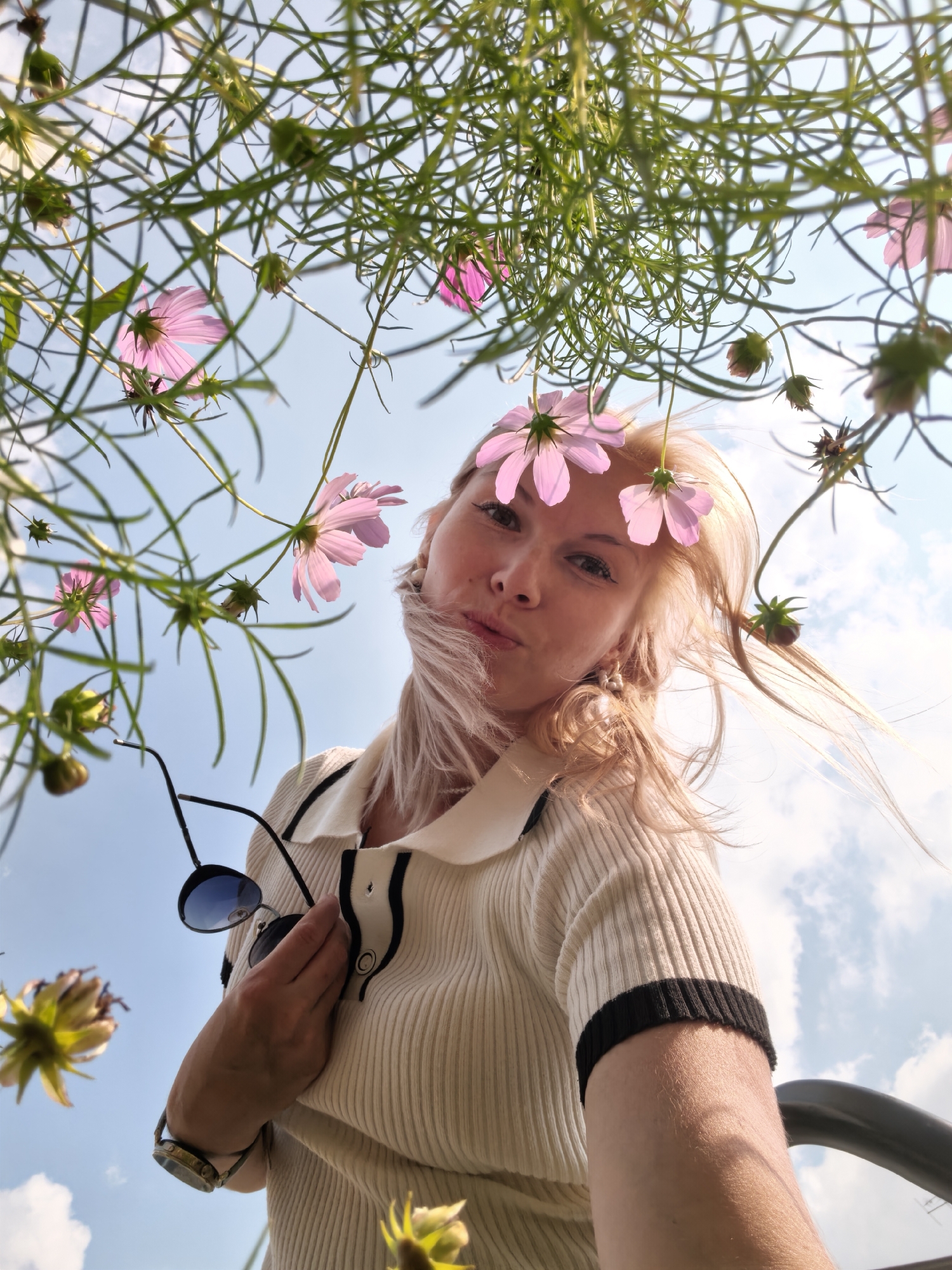 This is a low-angle, outdoor, full-shot photograph of a young Caucasian woman from the chest up. She has fair skin, blonde hair, and is looking directly at the camera. She is wearing a ribbed, cream-colored shirt with black trim on the collar and sleeves. She is holding a pair of dark sunglasses with blue lenses in her right hand, and a watch on her left wrist. The background consists of a blue sky with some white clouds, and a dense cluster of green plants with pink and yellow flowers. The image is taken at a downward angle, as if the viewer is looking up from the ground. The location of this photograph is not determinable from the image itself; therefore, it cannot be confirmed to be in Sterlitamak, Russia.