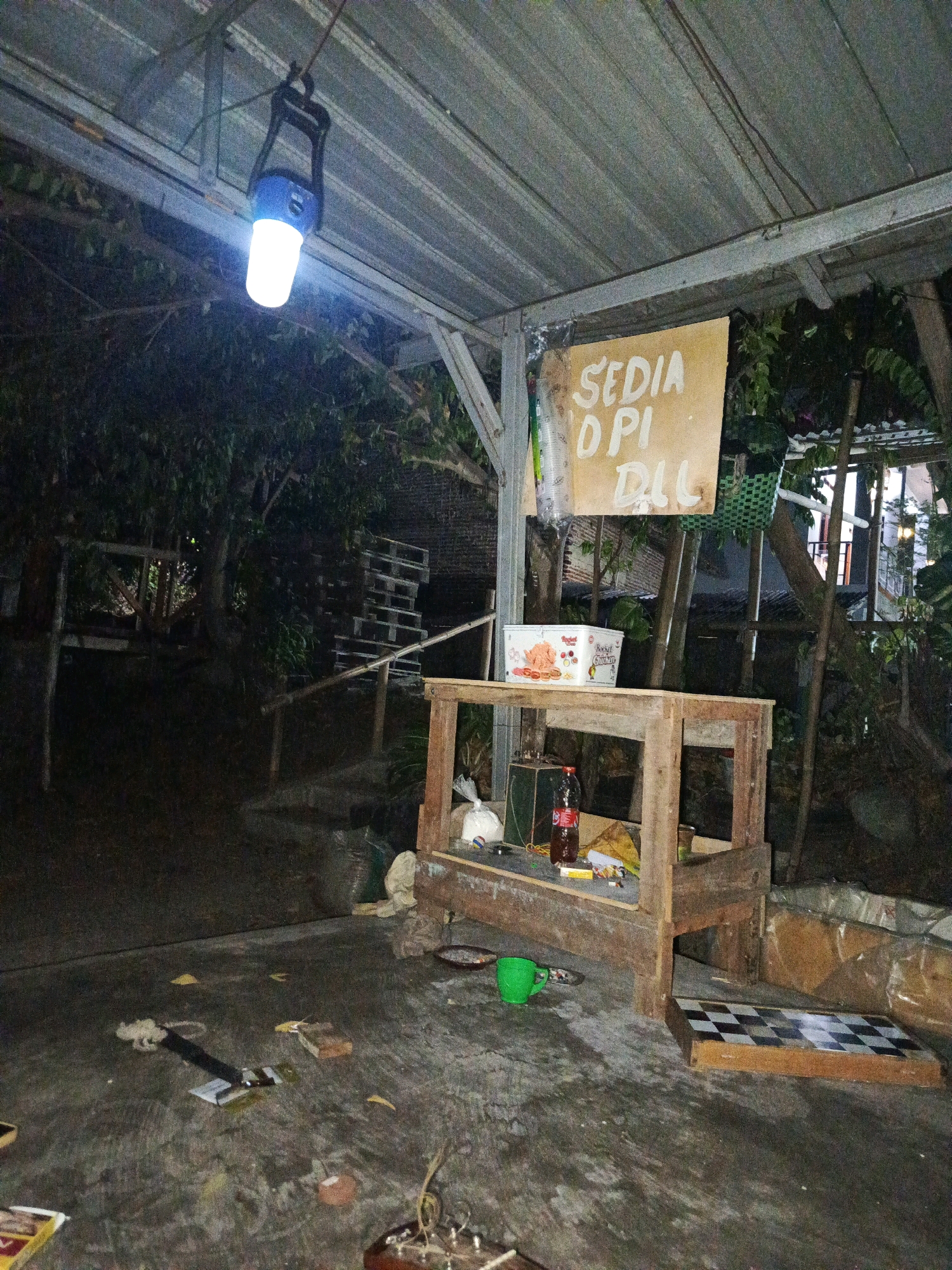 A dimly lit, outdoor covered area, possibly a stall or workshop, is captured at night in Tegal, Indonesia. A bright LED lantern hangs from the corrugated metal roof, illuminating a wooden stall in the foreground. On the stall, a bottle of soda, a small package, and a green mug are visible. A checkerboard rests on the ground next to the stall. The ground is concrete and cluttered with various debris, including a few yellow objects and what appears to be a fallen sign. In the background, stacks of wooden pallets and dense foliage suggest a somewhat rural or less developed urban setting. A sign with handwritten text in Indonesian, "SEDIA OPI DLL," which translates to "Serving Coffee Etc.," is posted on a wooden pole. The overall atmosphere is humble and functional.