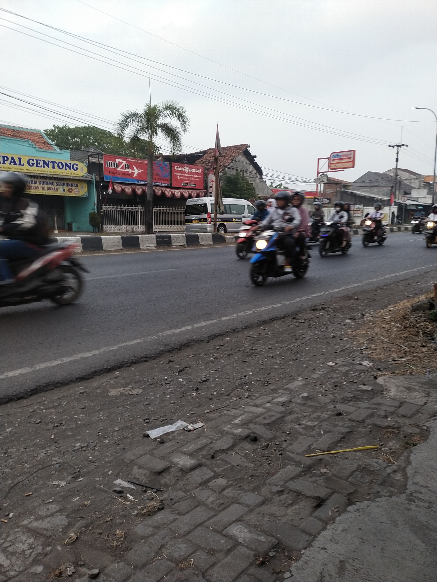 This image captures a street scene in Kemlakagede, Indonesia, on an overcast day. The foreground shows a paved road with a gravelly shoulder and a sidewalk constructed of interlocking bricks, littered with debris. Several motorcycles, some with riders, are in motion on the road. To the left, a shop with a turquoise facade displays signage for "MPAL GENTONG" and "AYAM GORENG SEBRONGAN". Next to it, a red banner advertises "MUZA TOUR & TRAVEL" and another banner, partially obscured, reads "LION PARCEL". Further down the street, a white van with "OBIBANS.CO.ID" branding is parked. In the background, buildings with red-tiled roofs are visible, along with power lines and a streetlamp. The sky is a uniform grey, suggesting cloudy weather.