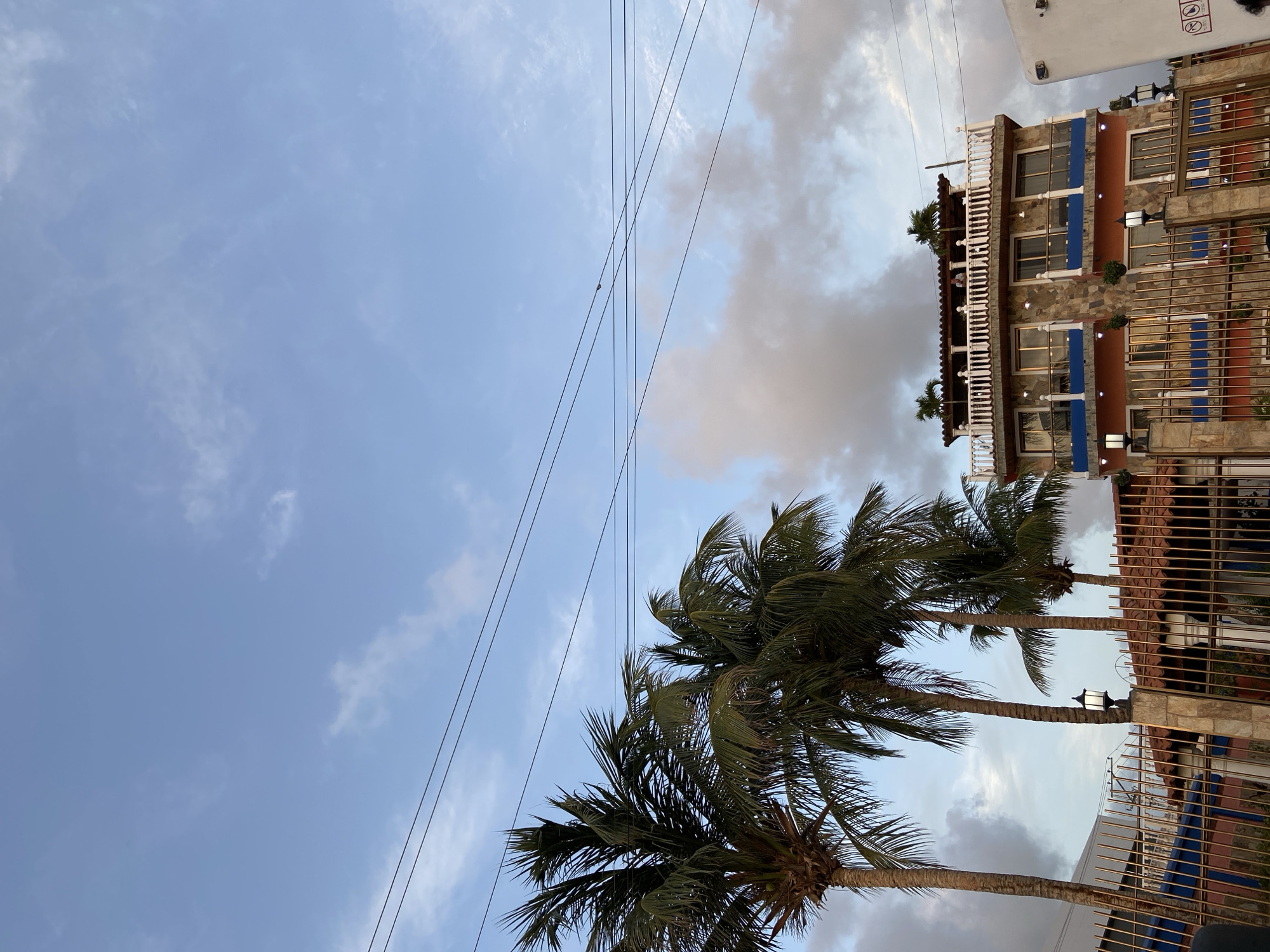 The image depicts a building with multiple stories, a balcony with white railings, and palm trees in the foreground. The building has an orange lower level with blue accents and stone-like siding on the upper levels. The sky is blue with scattered clouds. Power lines are visible across the sky. A metal fence separates the foreground from the building. The scene appears to be in Juan Griego, Venezuela.