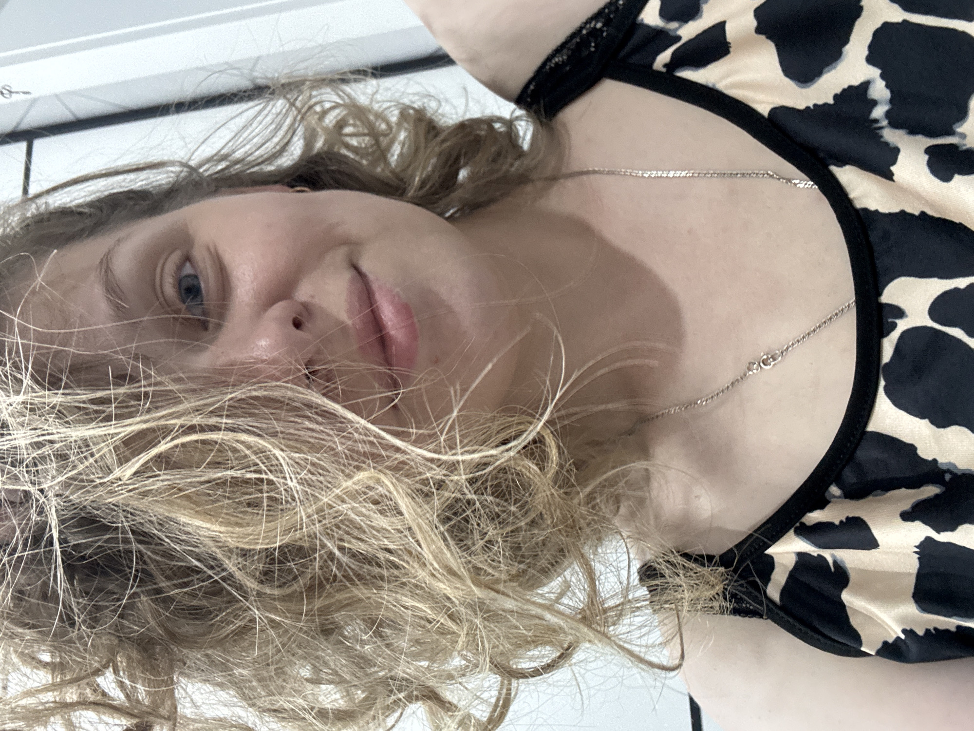 This is a close-up, overhead shot of a young woman lying down. She has fair skin, blonde curly hair that partially obscures her face, and blue eyes. She is wearing a black and beige patterned top with lace trim. A thin silver chain necklace is visible around her neck. The scene appears to be indoors, possibly a bedroom, with a white surface in the background. The lighting suggests it is daytime. The woman's expression is relaxed and she seems to be looking directly at the camera. There is no visible text in the image. The location context provided is Mikhaylovsk, Russia.