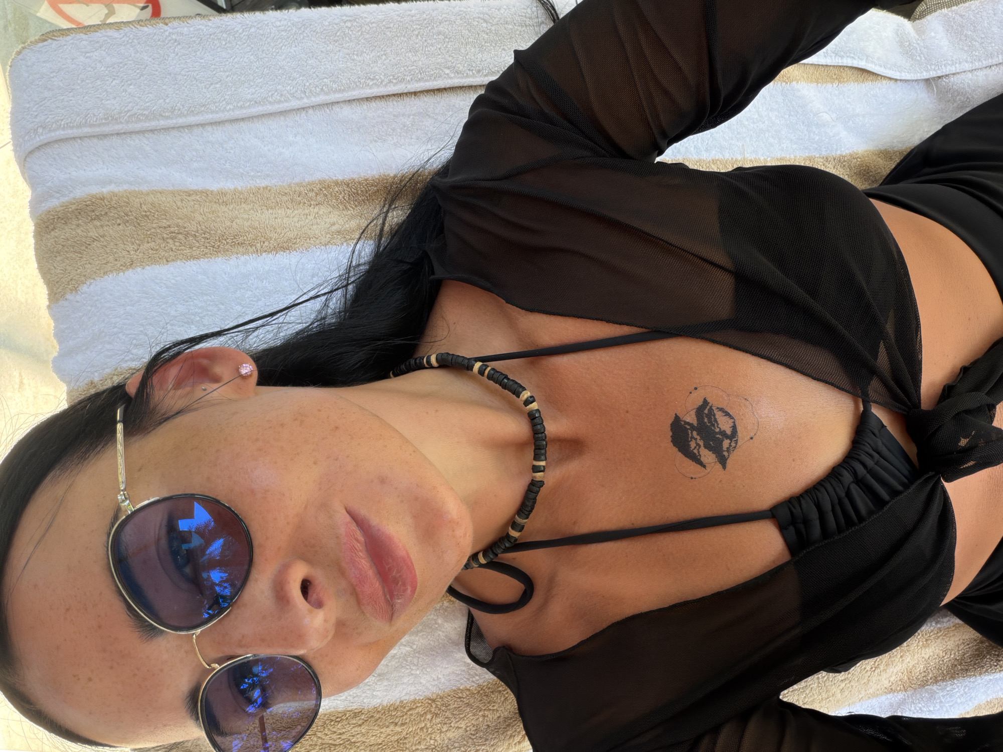 A woman is lying on a towel in Nusa Dua, Indonesia. She is wearing sunglasses, a black necklace, a black bikini, and a black mesh cover-up. The towel is white with beige stripes. The woman has a tattoo on her chest.