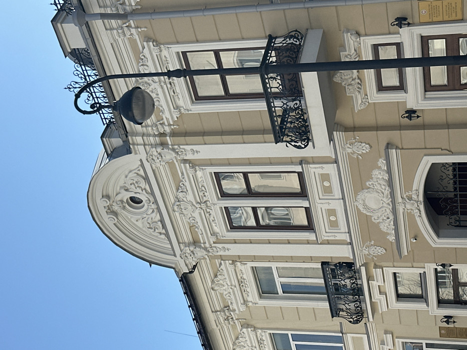 The image displays the ornate facade of a multi-story building, identified as the Ministry of Justice of the Republic of Tatarstan, ZAGS Administration, located at Kremlevskaya Street 12/20 in Kazan, Russia. The building features a light beige exterior adorned with extensive white stucco ornamentation in neoclassical and Baroque revival styles, including elaborate cornices, pilasters, scrolled motifs, and decorative wreaths.
The upper visible section of the building includes a prominent arched pediment with a central oculus (round window), surrounded by intricate white scrollwork and garlands. Below this, multiple rectangular windows with dark brown frames are visible; some contain white internal frames, and all exhibit reflections of the surrounding environment. Two black wrought-iron balconies with elaborate railings are present on the second visible story: one is a full cantilevered balcony, and the other is a smaller, false balcony situated beneath a window. A large circular stucco wreath decoration is prominently displayed on the facade below the main cantilevered balcony. Two wall-mounted lanterns and a small white security camera are visible on the lower right section of the building. A brown rectangular sign with white text in Russian and Tatar, identifying the Ministry of Justice of the Republic of Tatarstan, ZAGS Administration, is affixed to the lower right facade.
In the foreground, a tall, black, ornate street lamp post with a spherical globe light fixture stands, partially obscuring the right side of the building's facade. A thin, dark spike or antenna protrudes from the roofline on the left. The scene is illuminated by natural daylight under a clear, bright blue sky. No individuals, actions, or interactions are present.