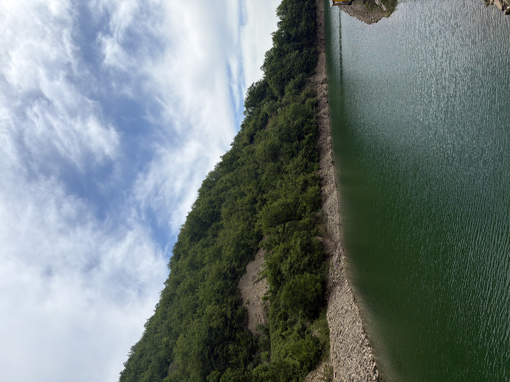 The image shows a verdant hillside leading down to a body of water under a partly cloudy sky. The hillside is covered in dense green foliage. The water, possibly a lake or reservoir, has a dark green hue and a slight ripple on the surface. A narrow strip of rocky terrain forms the shoreline. The sky is blue with streaks of white clouds. The location is likely near the city of Gračac, Bosnia and Herzegovina.