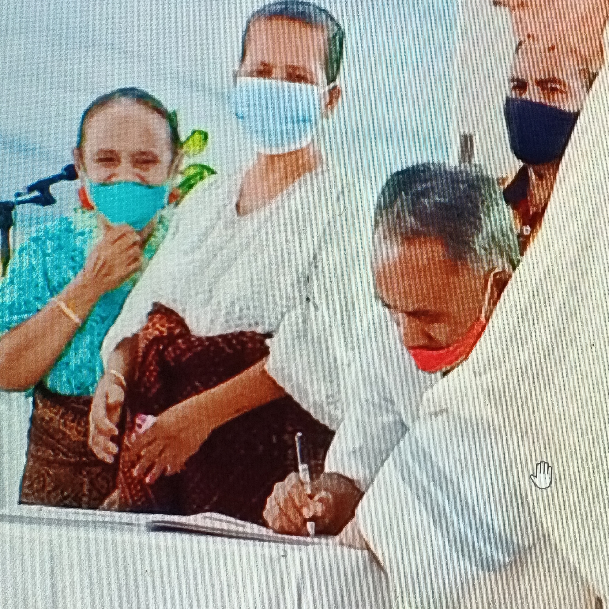 The image captures a scene at an event in Ende, Indonesia, possibly a formal signing ceremony. In the foreground, a man, wearing a white collared shirt and a red mask, is seated and actively signing a document with a pen. To his left, a woman in a blue patterned top and a patterned skirt, also wearing a turquoise mask and a bracelet, is looking towards the left of the frame. Behind her, another woman, wearing a white lace top and a patterned skirt, with a light blue mask, is also looking to the left. Further to the right, a man wearing a dark blue mask is partially visible. The setting appears to be indoors or under a covered area, suggested by the backdrop of a textured surface and a table in the foreground where the signing is taking place.