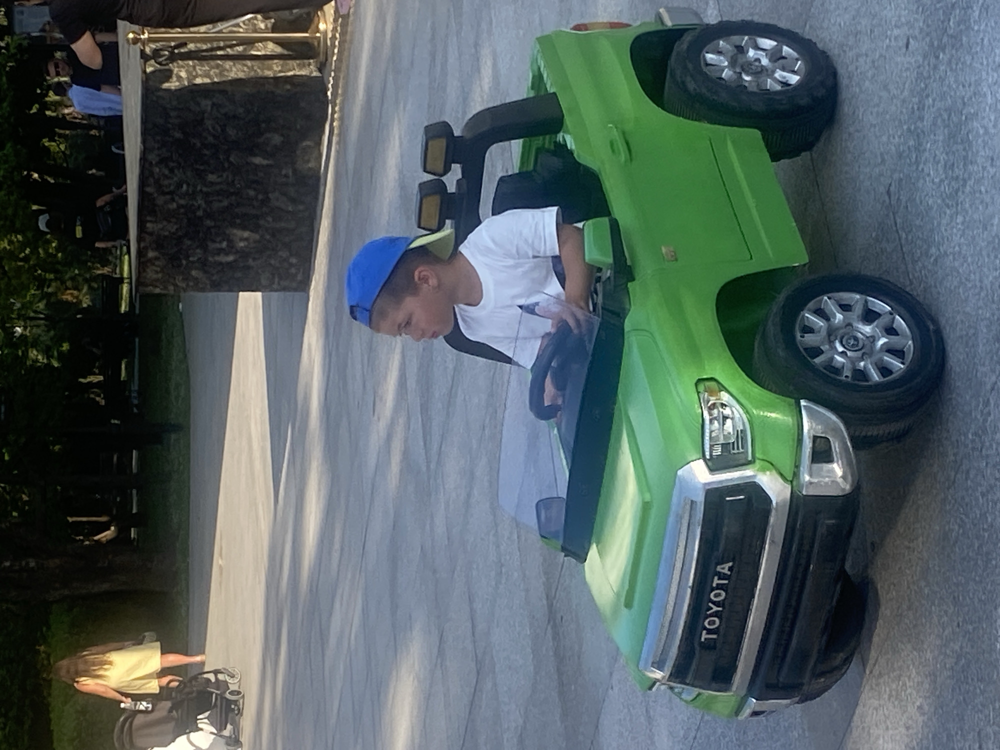 A young boy wearing a blue baseball cap and a white t-shirt is sitting in the driver's seat of a bright green toy electric truck. The truck is a replica of a Toyota Tundra, complete with a realistic grill and headlights. The boy is looking to his left, with his hands on the steering wheel. He appears to be in an outdoor public space, possibly a park or plaza, with paved ground and trees in the background. The lighting suggests it is daytime, with sunlight casting shadows on the ground. In the background, other people can be seen, some standing and others walking. One person is pushing a stroller. The environment appears to be well-maintained, with the paved area suggesting a developed urban or recreational area. Based on the context provided, this scene is taking place in the city of Sochi, Russia.