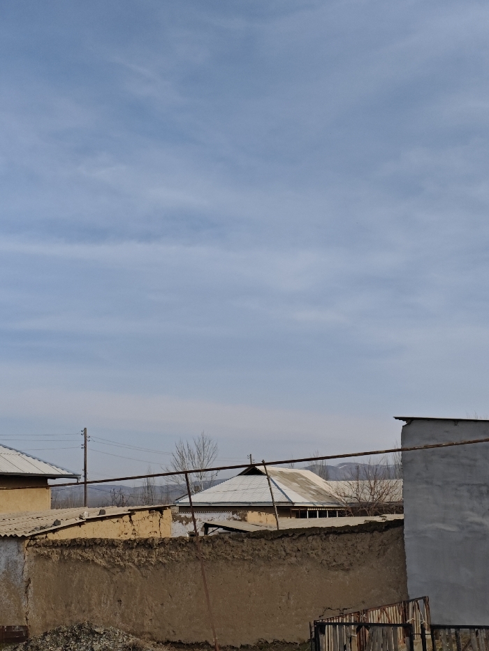 The image displays an outdoor, daytime scene in Abdukarim, Uzbekistan, featuring various structures under a light blue, overcast sky. The foreground is dominated by a weathered, light brown mud or adobe wall, approximately 1.5 meters in height, extending from the lower-left to the lower-right. A pile of dark, broken material lies at the base of this wall on the left. Partially visible behind the lower-right section of the wall is a rusty, dark-colored metal gate or fence. Several rusty metal bars or pipes traverse horizontally across the mid-ground, with one prominent bar running diagonally from mid-left to mid-right, passing over the mud wall.
In the mid-ground, multiple residential or ancillary buildings are visible. On the far left, a light yellow-walled structure with a light-colored, pitched roof is partially shown. Further to the right, behind the mud wall, is another building with a light-colored, corrugated metal pitched roof. To its right stands a larger structure with white or light-grey walls and a similar corrugated metal pitched roof, featuring a band of small, rectangular windows directly below the roofline. On the far right, a tall, smooth, grey-painted wall of a building with an unseen or flat roof extends to the frame's edge. A dark utility pole with multiple horizontal crossarms and wires is positioned in the mid-left background.
The background reveals scattered leafless trees, indicating a dormant season. Distant, hazy blue-grey mountains or hills are visible along the horizon line. No individuals, animals, or vehicles are present in the depicted scene.