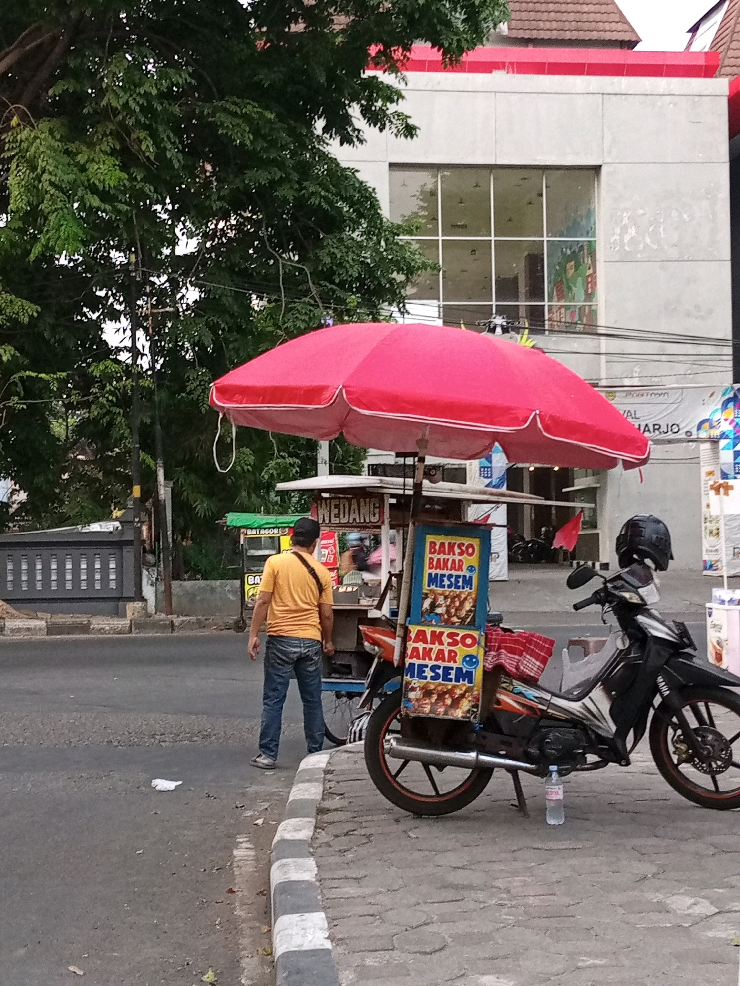 A street food vendor is parked on the side of a road in Sukoharjo, Indonesia. The vendor is operating from a cart topped with a large red umbrella, with a motorcycle parked beside it. The cart displays advertisements for "Bakso Bakar Mesem" (Grilled Smiling Meatballs). A man in a yellow shirt and blue jeans stands near the cart, facing away from the camera. The street is paved with asphalt and bordered by a sidewalk made of cobblestones. In the background, a modern building with large windows stands behind a lush green tree. Electrical wires and poles are visible in the background.