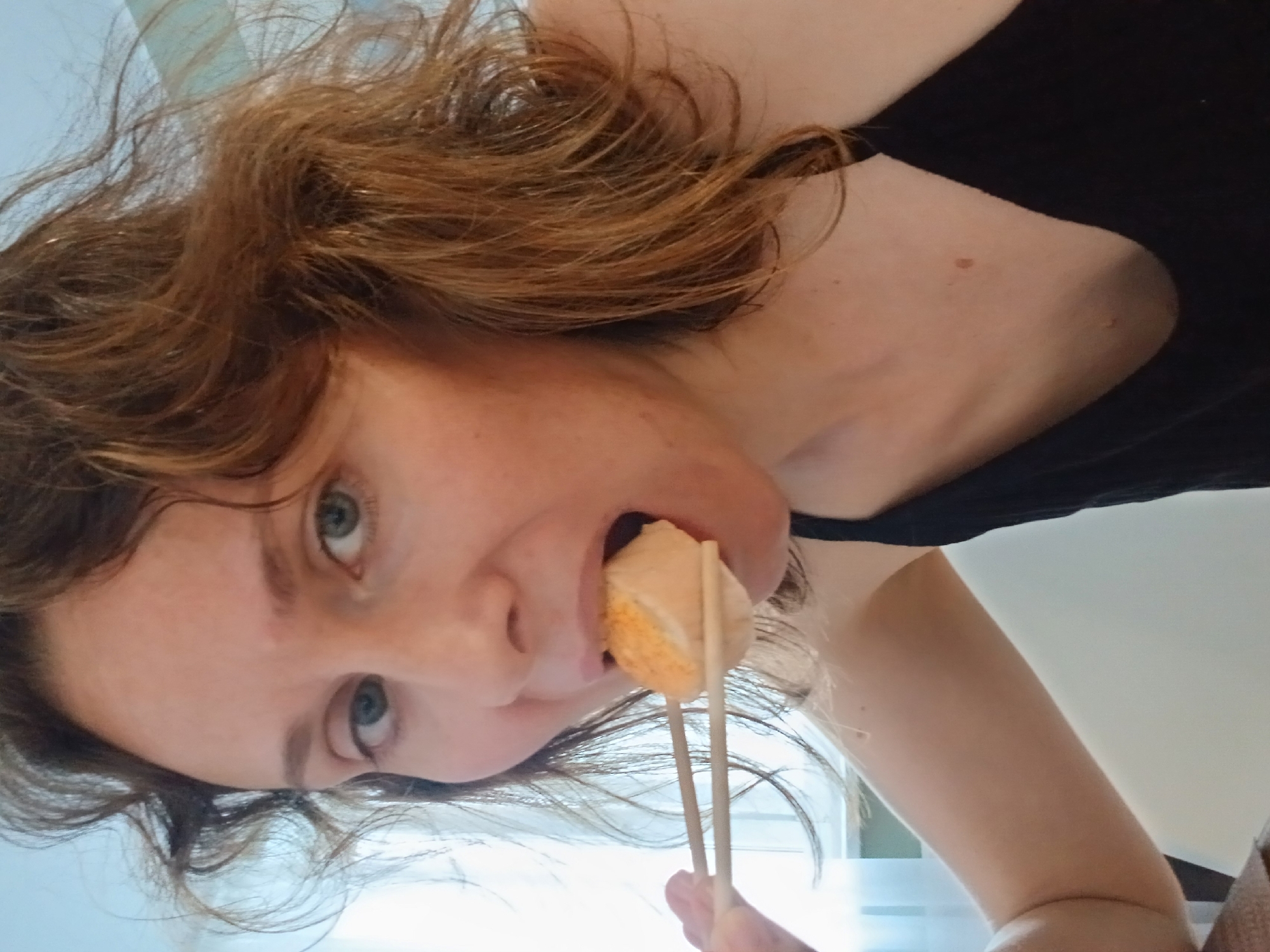 A medium close-up shot captures a woman with auburn hair, facing forward and partially eating a piece of food held by chopsticks. She is wearing a black camisole. The background is blurred, with hints of a window and blinds suggesting an indoor setting. The lighting appears to be natural, coming from the left side of the frame.