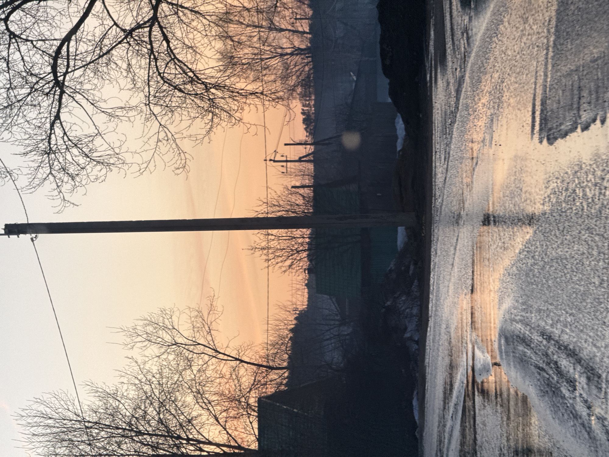 The image depicts a roadside scene at twilight. A wooden utility pole with electrical wires runs vertically in the middle of the frame. Leafless tree branches are silhouetted against the sky, which is a gradient of light orange to gray. The road surface appears wet and reflects the colors of the sky. A blurry section of a building is visible in the background. Based on the information provided, it is located in Chaikovsky, Russia.