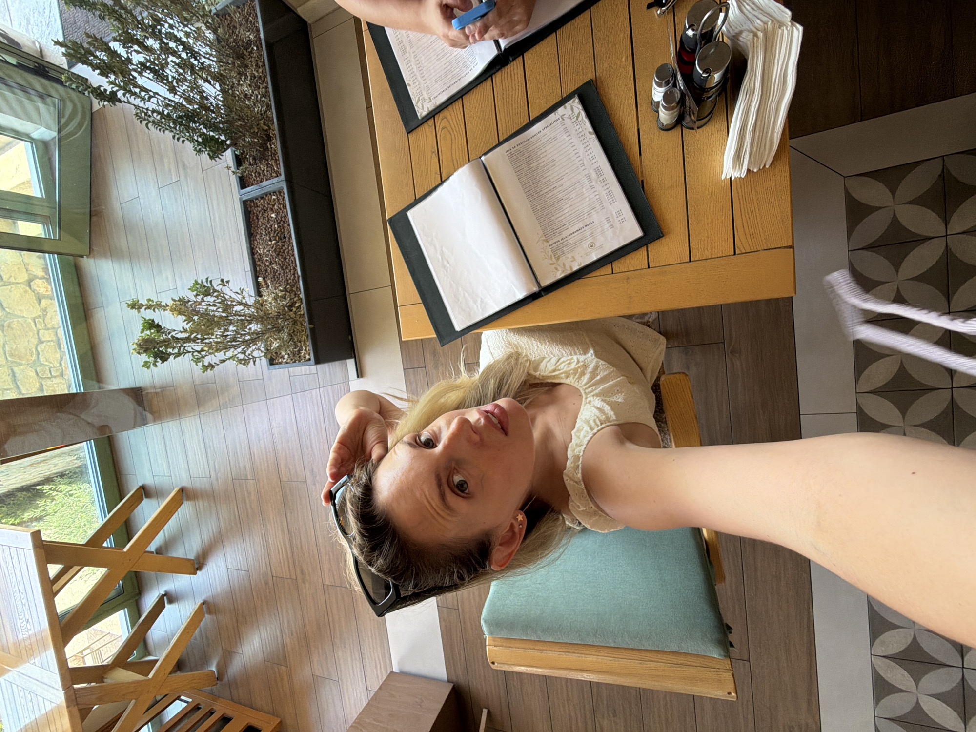 A woman is sitting at a wooden table in what appears to be an outdoor seating area of a restaurant, possibly in Bigor, Montenegro. She is taking a selfie with her arm extended, looking directly at the camera with an open mouth and raised eyebrows. Her left hand is resting on her head, and sunglasses are perched on top of her blonde hair. She is wearing a light-colored, ruffled blouse.

In front of her on the table are two menus in black binders, one open to display its pages, and the other partially obscured by a person's hand holding a pen. A stack of white napkins is placed to the right of the menus, next to a holder containing salt and pepper shakers and possibly a corkscrew.

The table is made of light-colored wood with a slatted surface. In the background, there are more wooden tables and chairs, a large window with a view of greenery, and planters with dried plants. The flooring is a combination of wooden planks and patterned tiles. The lighting suggests it is daytime, with sunlight filtering into the area.