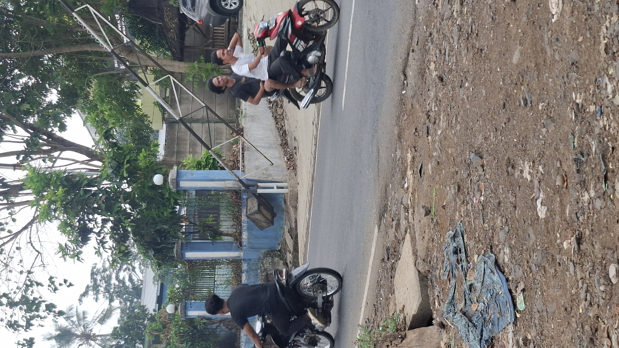 In Cilaku, Indonesia, a dirt and gravel roadside is littered with debris. A blue and white fence runs along the property line. A black motorcycle with a single rider wearing a black shirt and pants is seen in motion on the asphalt road. Further down the road, a red motorcycle carries two passengers, both men. The man in front wears a white t-shirt and black pants, while the man behind him wears a black t-shirt and black pants. They are also in motion on the road. In the background, a large metal structure, possibly part of a construction or utility pole, leans precariously, supported by angled beams. Trees and vegetation are visible behind the fence. A dark-colored car is parked on the right side of the image. The sky is overcast.