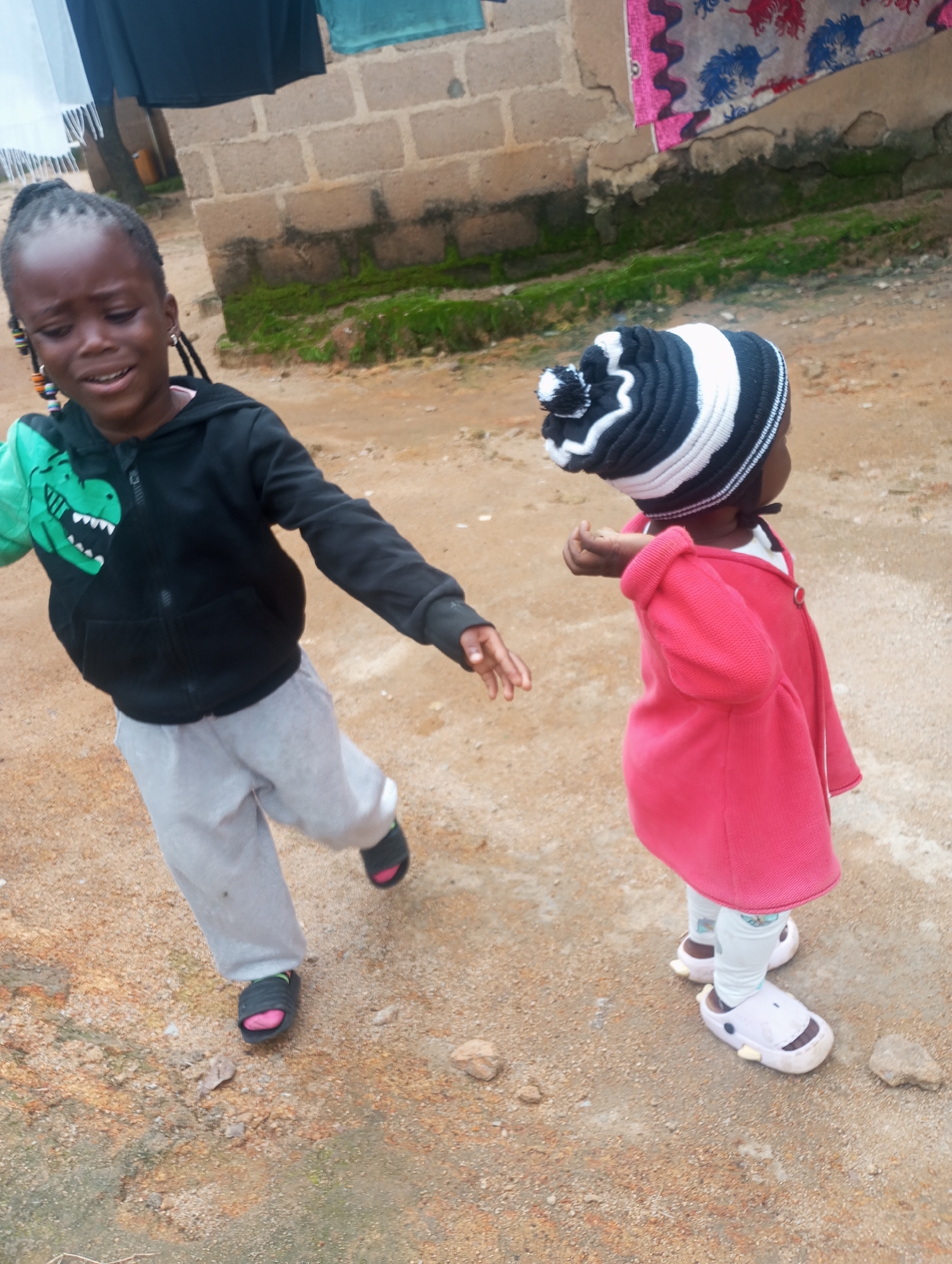 Two young children are outside on a dirt path in front of a rough brick building. The building appears to be partially covered in moss and has laundry hanging on a line in front of it. The child on the left, wearing a black hoodie with a green dinosaur on it and light gray pants, is looking forward with their mouth open, appearing to be crying or yelling. This child has dark braided hair with colorful beads. The child on the right is wearing a pink coat, white leggings, and a black and white striped hat with a pom-pom. This child is facing away from the camera. The ground is composed of dirt and small rocks, with some patchy green growth visible in the background. The scene is likely taking place in a residential area of Jos, Nigeria, given the visual context.