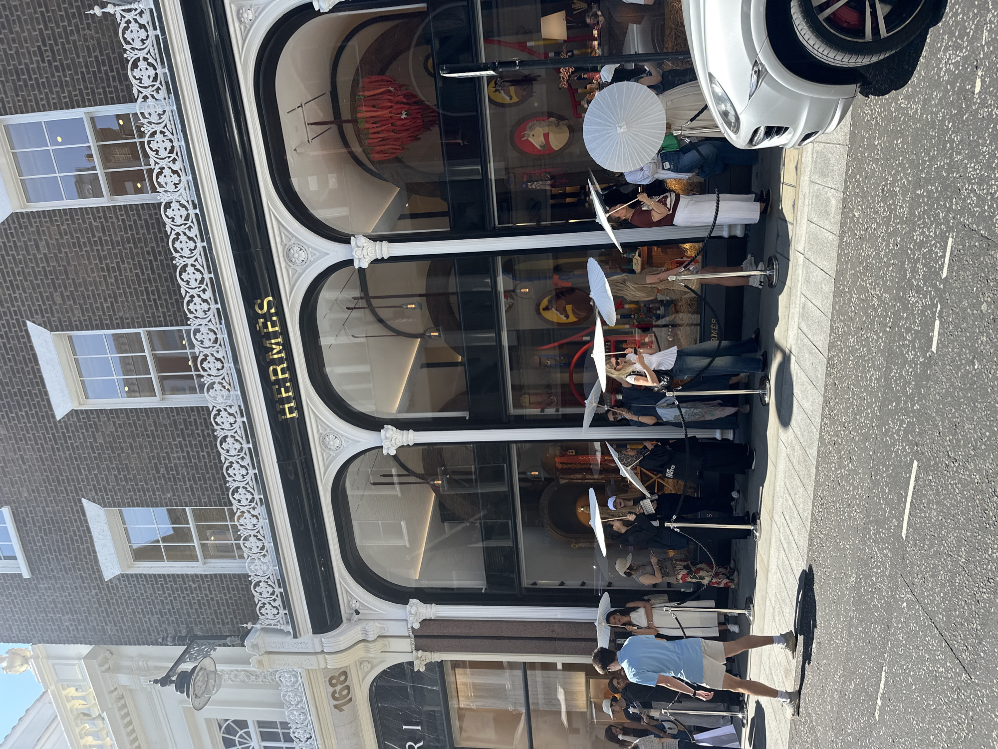The image shows the exterior of a luxury retail store, identified by the prominent "HERMÈS" signage in gold lettering above arched display windows. The building is located in a city, with architecture that suggests a European setting. Specifically, the presence of the number "168" on an adjacent building, along with the style of the storefronts, points to a location in London, United Kingdom.
A line of people is formed outside the Hermès store, with many holding white parasols, suggesting sunny weather. The people are dressed in casual to semi-formal attire. Some individuals are looking into the store windows, while others appear to be waiting. The street in front of the store is paved with asphalt and has white line markings. A silver SUV is parked on the right side of the frame, partially obscuring the view of the sidewalk and street. The storefront itself features large glass windows showcasing merchandise, including what appears to be a chandelier-like display in one window and framed images of horses in another. The building's facade is detailed with decorative white architectural elements, including ornate railings above the windows and classical columns framing the arched entrances. The overall scene depicts a bustling urban environment with a focus on high-end retail.