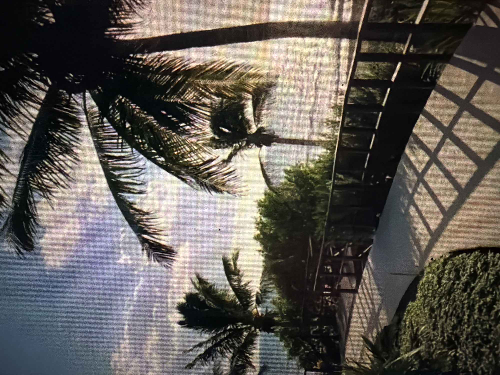 The image depicts a tropical pathway leading towards a body of water, likely the ocean. Several palm trees are visible, with fronds silhouetted against a partly cloudy sky. A wooden railing lines one side of the pathway, which is paved with a light-colored material. The water reflects the sunlight, creating a shimmering effect. The scene appears to be set in Baan Mae Nam, Thailand, given the presence of palm trees and proximity to water, characteristic of coastal areas in the region. The overall atmosphere is serene and summery.