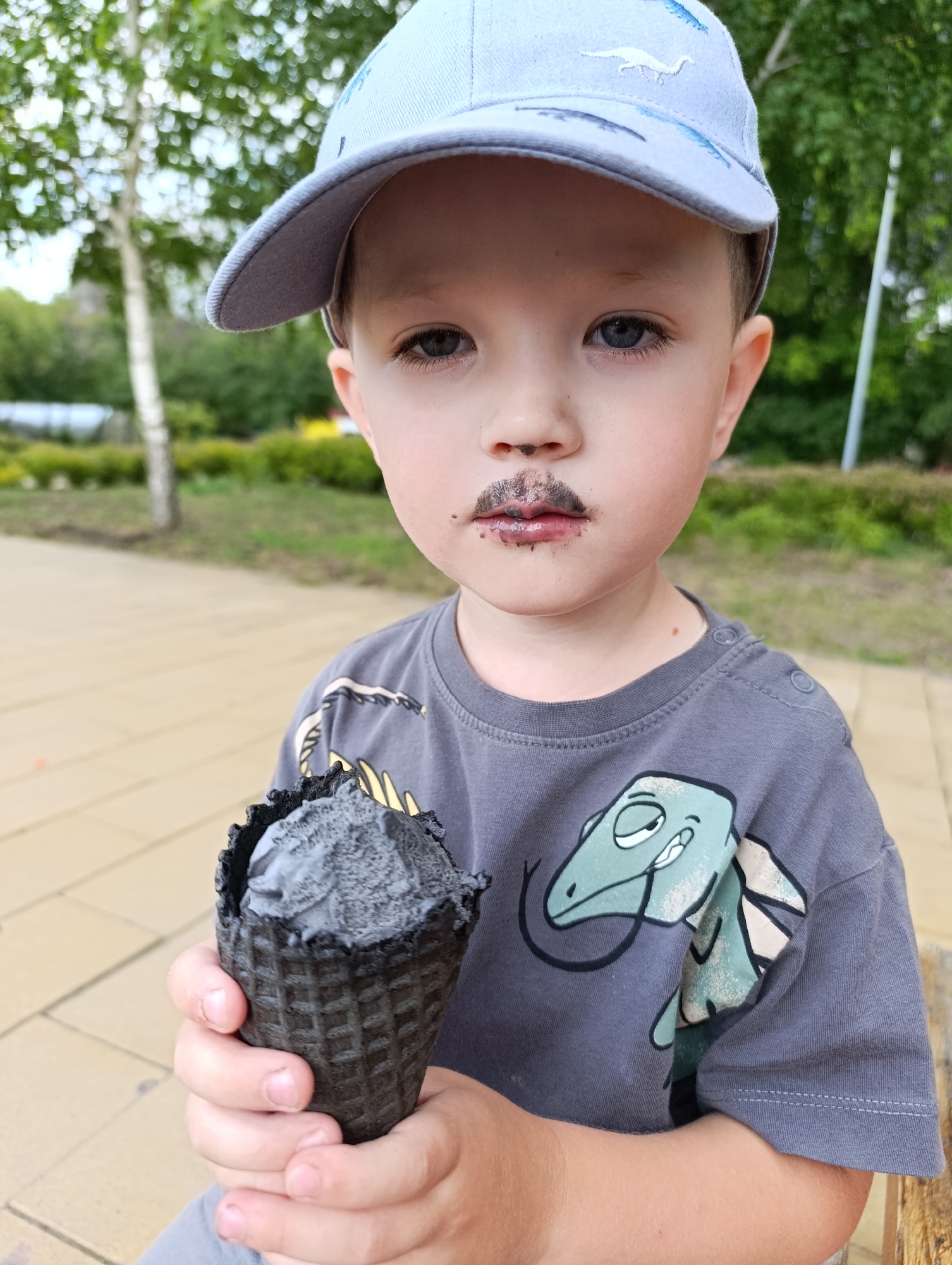 A close-up shot captures a young Caucasian boy wearing a light blue baseball cap and a gray t-shirt with a cartoon dinosaur print. He holds a black ice cream cone in his right hand. The boy's upper lip is smudged with what appears to be chocolate ice cream, resembling a mustache. He looks directly at the camera with a neutral expression. The background is softly blurred, showing green foliage, trees, and a light brown paved area, suggesting an outdoor setting such as a park or a public space within the city of Novokuybyshevsk, Russia.