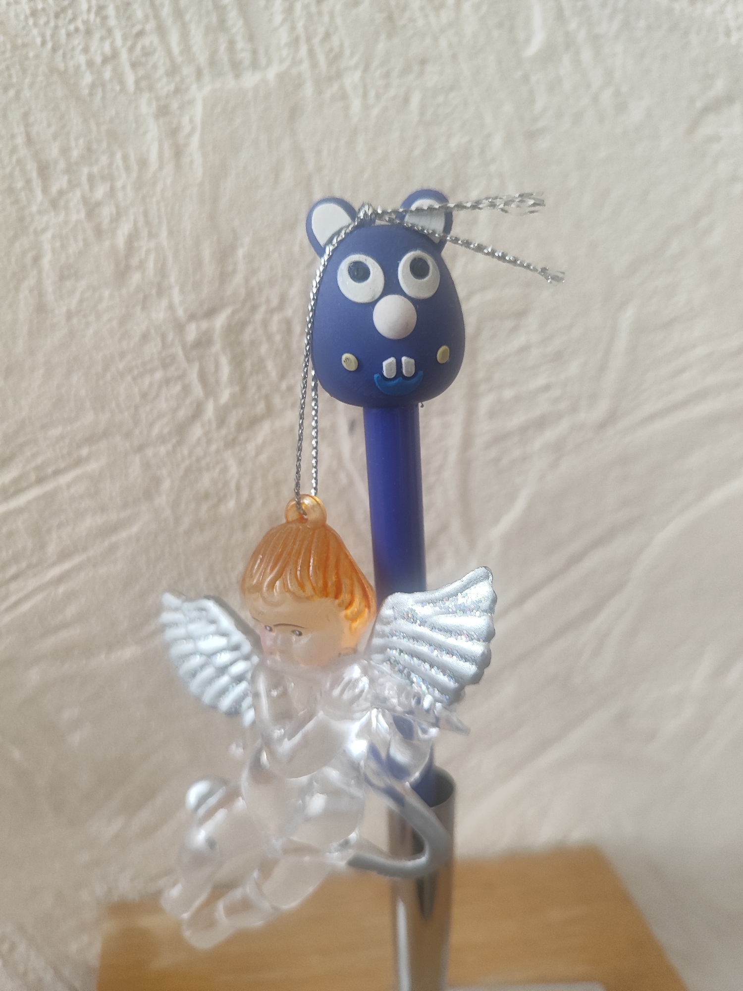 A close-up shot shows a blue pen with a playful, cartoonish design at the top, resembling a bear with big eyes and a small mouth. A silver chain hangs from the top of the pen, suspending a small, translucent angel figurine. The angel has light orange hair and glittery silver wings. The pen and angel are positioned in the foreground, with a textured, light-colored surface serving as the background. The lighting suggests an indoor setting, likely with natural light. No text is visible in the image.