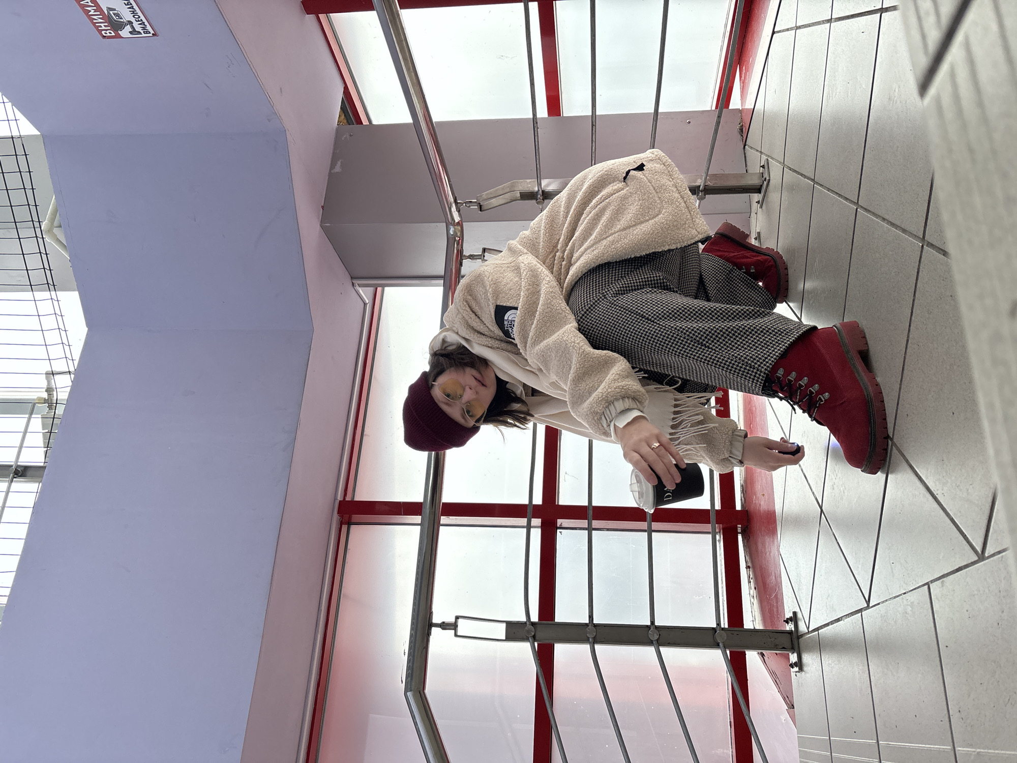 A person wearing a red beret, yellow-tinted glasses, a cream-colored teddy jacket, plaid pants, and red boots is sitting on the stairs of a building in Chaikovsky, Russia, holding a black coffee cup. The person is positioned between the gray metal railings of the staircase, against a backdrop of red window frames and white walls. The stairs have white and gray tiled flooring. A sign in Russian is partially visible on the wall above.