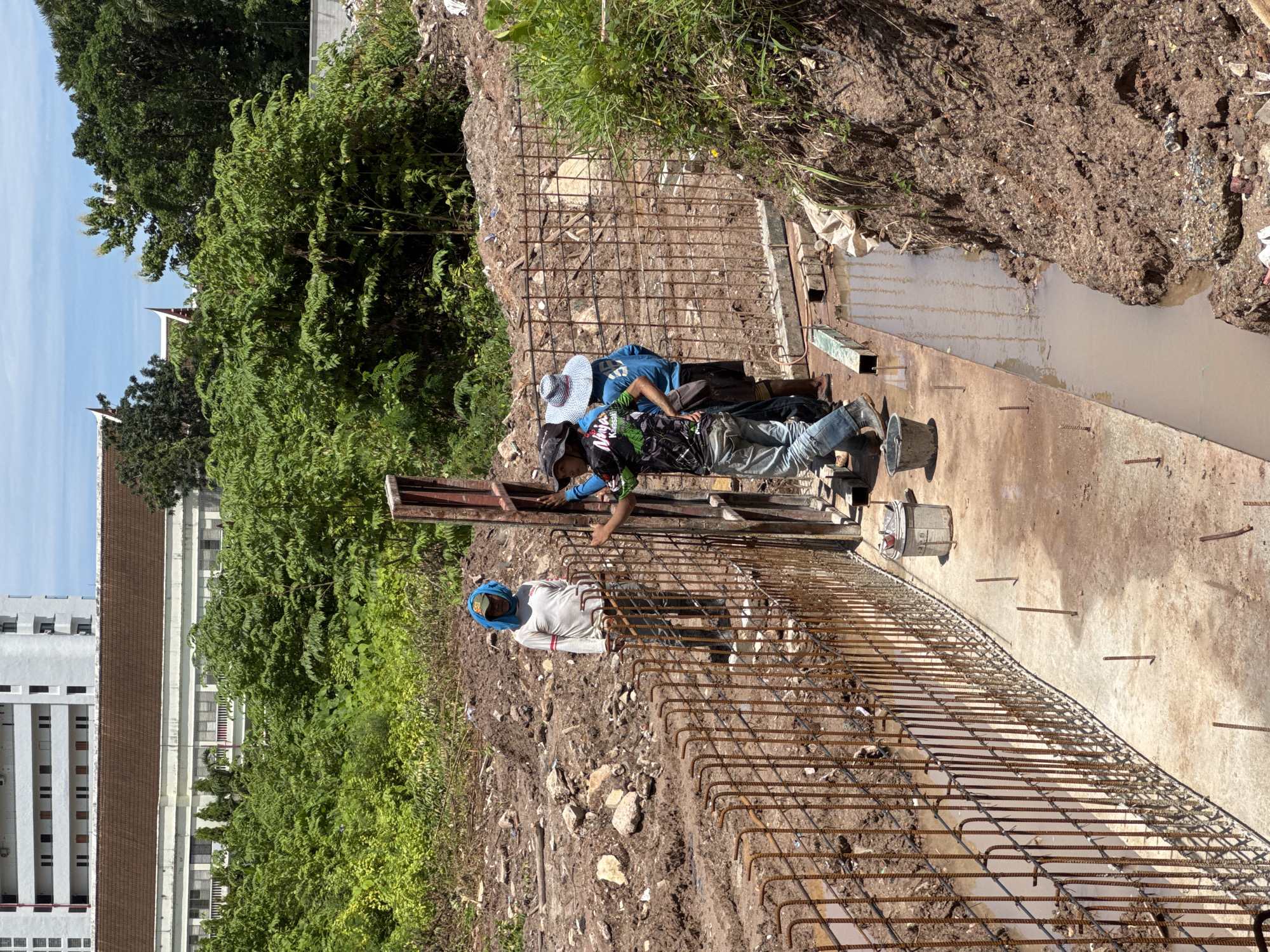 Three construction workers are visible at a construction site in Patong, Thailand. The foreground features a concrete foundation with rebar protruding from it, forming a wall. A narrow trench filled with muddy water runs alongside the foundation. In the mid-ground, the workers are positioned near the rebar structure, appearing to be engaged in construction activities. One worker, wearing a blue head covering and a white long-sleeved shirt, stands observing. Another worker, in a blue shirt and a straw hat, is partly obscured. The third worker, wearing a black t-shirt with a graphic design and a hat, is actively working on the rebar. The background consists of a steep embankment of dirt and vegetation, with buildings visible in the distance. The sky is clear and blue. The scene suggests ongoing construction work in an outdoor environment.