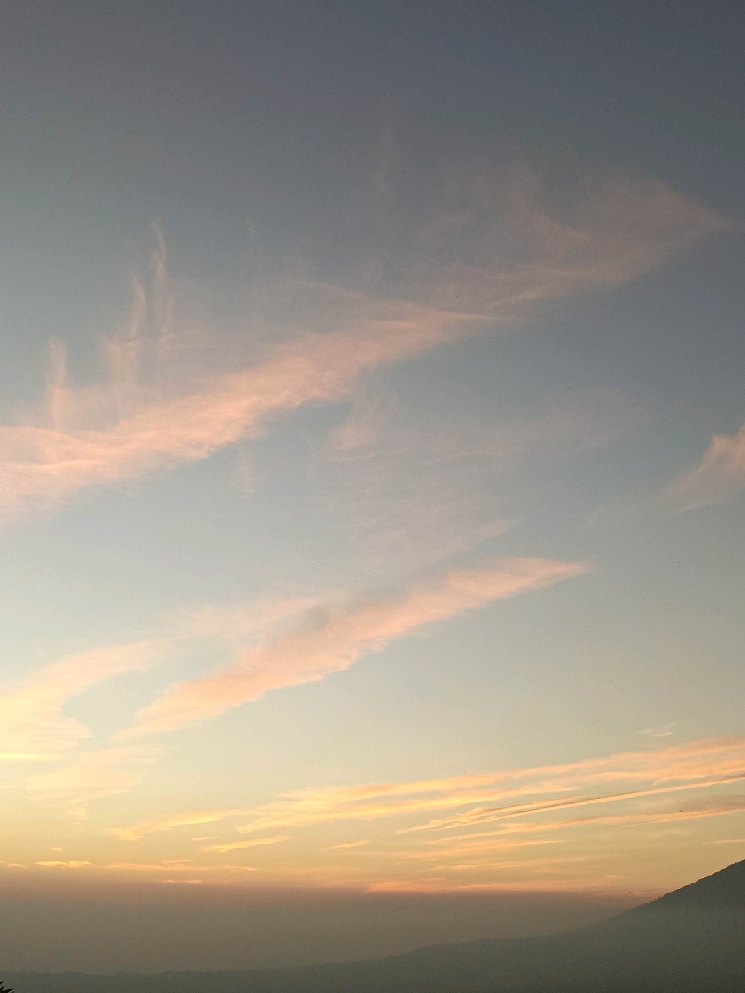 This image depicts a hazy sunset over a silhouetted landscape, possibly the outskirts of Butuh Kidul, Indonesia. The sky is a gradient of pale blue to soft orange, with wispy, pink-tinged clouds scattered throughout. The lower portion of the image is dominated by a dark, indistinct landmass, suggesting hills or mountains shrouded in atmospheric haze. The overall impression is one of a peaceful, quiet evening. There are no discernible people, specific objects, or actions visible in the scene.