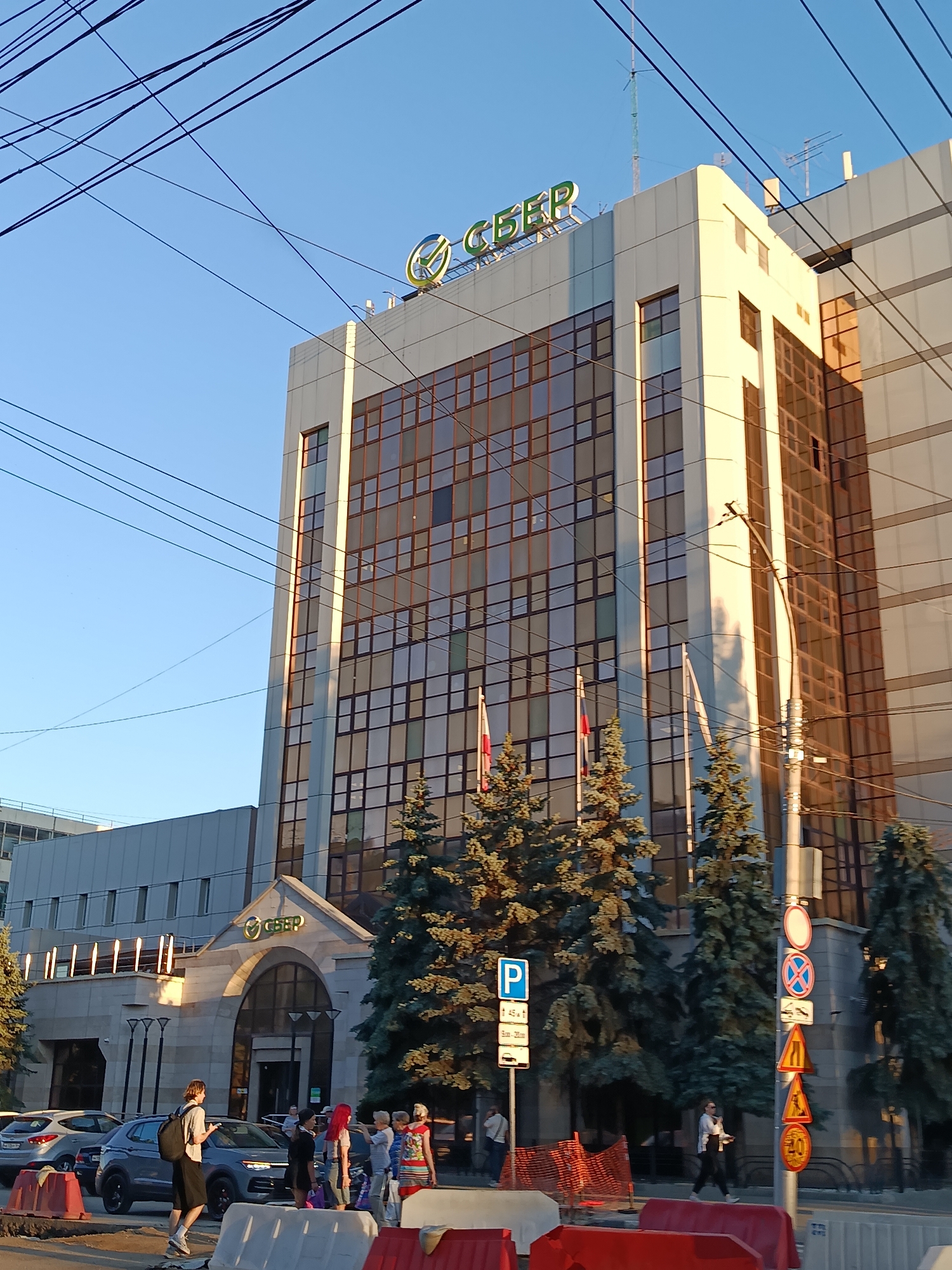 This media file depicts a street scene in Saratov, Russia, on a clear, sunny day with the golden hour light casting long shadows. The main subject is a modern office building with a prominent "СБЕР" (Sber) logo displayed on its facade. The building features large, reflective windows and a light-colored exterior.
In the foreground, several people are visible. A young man with a backpack walks towards the building, while a group of people, including a woman with red-dyed hair, are gathered near the entrance. A few cars are parked on the street, and construction barriers are present, suggesting ongoing work or traffic management.
Several traffic signs are mounted on a pole on the right side of the image, including parking signs, prohibition signs, and speed limit signs. Tall evergreen trees are planted in front of the building, adding a touch of nature to the urban landscape. The presence of power lines crisscrossing the sky is a common sight in urban environments. The overall atmosphere appears to be calm and ordinary, with people going about their day. The time of day is likely late afternoon or early evening, given the warm lighting and shadows.
