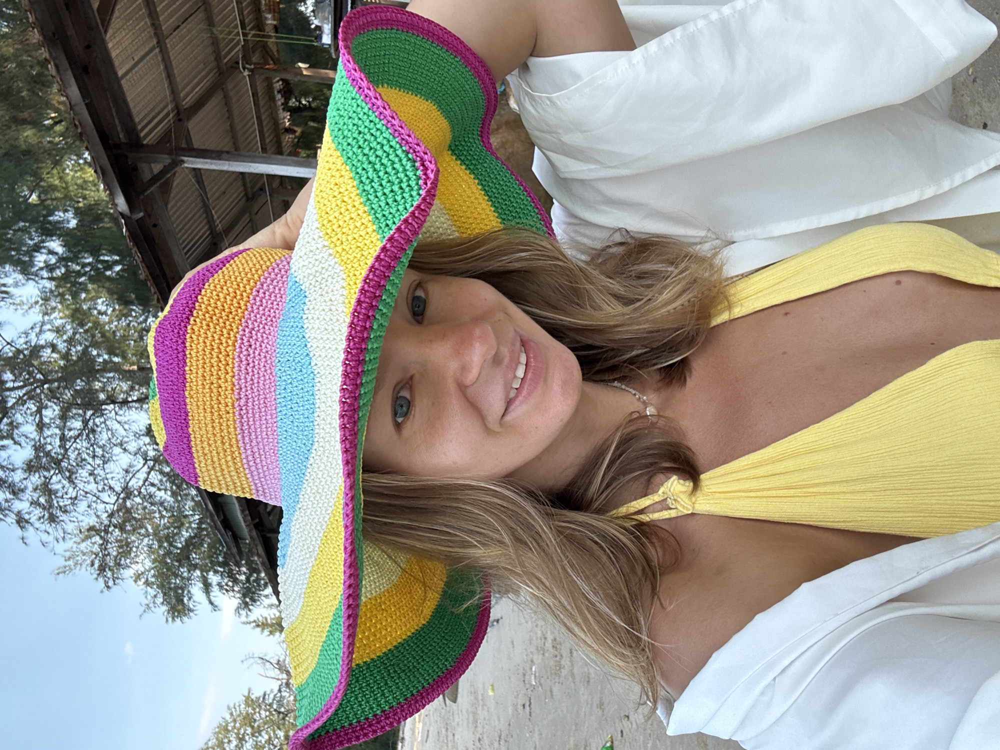 A woman is posing for a selfie on a beach. She is wearing a yellow bikini top and a white shirt draped over her shoulders. She has on a large, colorful crocheted sun hat with stripes of pink, orange, yellow, light blue, white, and green. The woman has blonde hair and is smiling at the camera. In the background, there is a sandy beach, trees, and a rustic wooden structure with a corrugated metal roof. The scene appears to be outdoors, possibly in a tropical location. There is no indication that this location is in Thuận Thành, Vietnam.