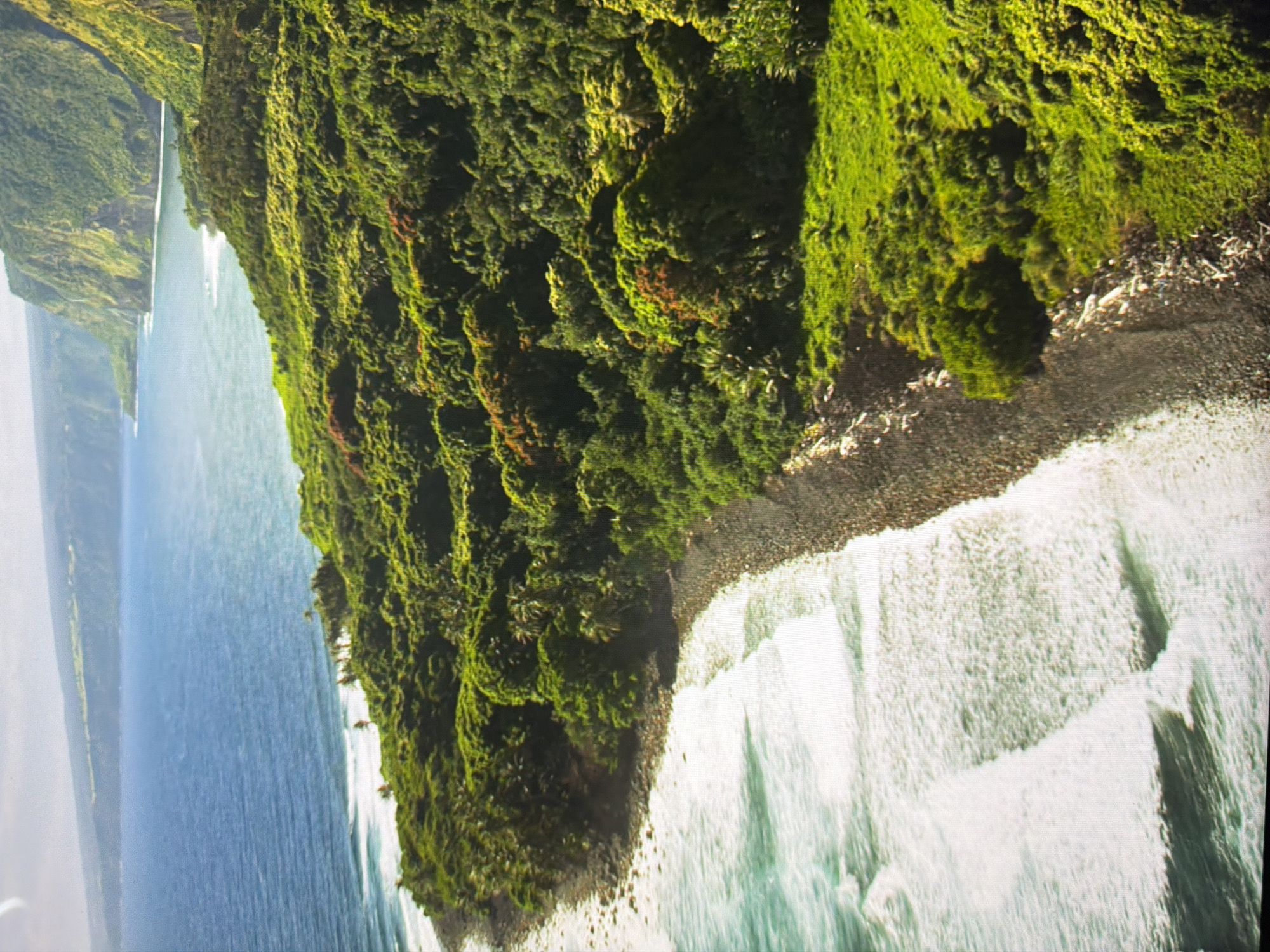 This is an aerial, high-angle, full shot of a lush, green, tropical coastline. The foreground features a dark, rocky beach with white waves crashing onto it. The ocean is a deep blue, with lighter turquoise hues closer to the shore. Beyond the beach, the land rises sharply, covered in dense, vibrant green vegetation, including palm trees and patches of reddish foliage. In the distance, imposing, verdant cliffs rise dramatically from the sea, their slopes covered in vegetation. The sky is a pale, hazy blue with some clouds. There are no people or man-made structures visible in the image. The scene depicts a remote and natural coastal landscape.