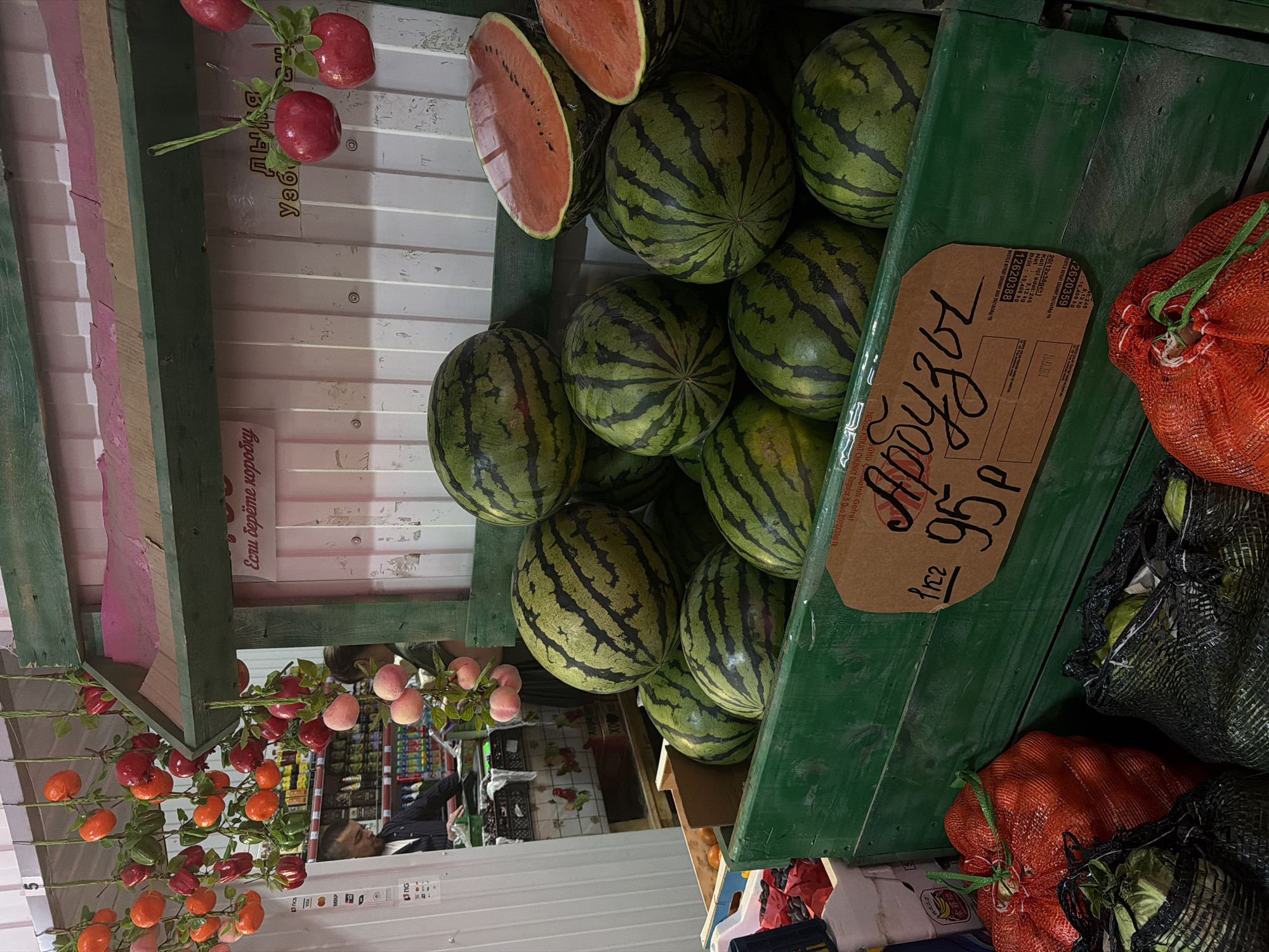 This is a close-up, eye-level shot of a fruit stall in Birobidzhan, Russia. The focal point is a green wooden crate filled with watermelons. Several whole watermelons, characterized by their dark green rinds with lighter green stripes, are visible. Two halves of a watermelon, revealing their bright red flesh, are also present. A price tag, handwritten in black ink on a light brown card, is affixed to the front of the crate, displaying the Cyrillic word "Арбузы" (Arbuzы - Watermelons) and the price "95 Р" (95 rubles).
Above the watermelons, hanging from green strings, are decorative items that resemble pomegranates and other red fruits, interspersed with green leaves. In the background, a white wall with vertical corrugated panels is visible. A section of the stall structure, painted green and pink, is also present.
To the left of the main fruit display, a man in a dark jacket and a person partially obscured are interacting at a counter, which appears to be a point of sale. Shelves stocked with various products, possibly food items, are visible behind them. Bags of produce, including a bright red mesh bag filled with round objects (likely tomatoes or peppers) and black mesh bags containing green items (possibly cabbage or lettuce), are positioned in the foreground to the left and right of the watermelon crate. The overall scene depicts a vibrant, open-air market setting.