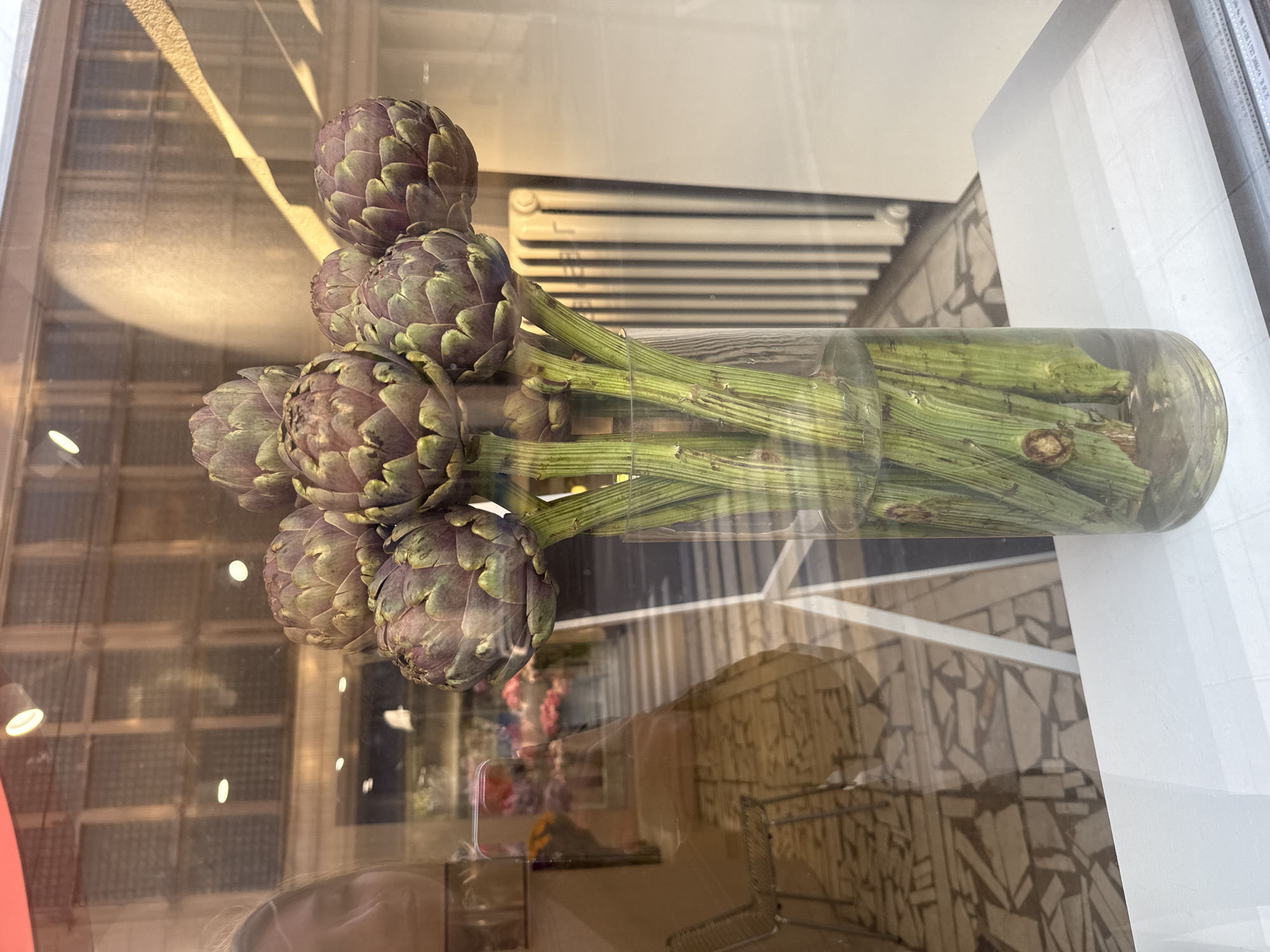 A still-life arrangement of artichokes in a glass vase is visible in a storefront window, likely in Saint Petersburg, Russia, as the radiator depicted is commonly used in Saint Petersburg, Russia. Nine artichokes are arranged in the vase, their purple-green heads prominent. The stems are long and green, bunched together and submerged in water within the cylindrical glass vase. The display is positioned against the backdrop of a window with a reflection of the surroundings. Details such as the building's architecture, mosaic tiled ground and the arrangement of items indicate the scene is likely in a shop or boutique setting in Saint Petersburg, Russia.
