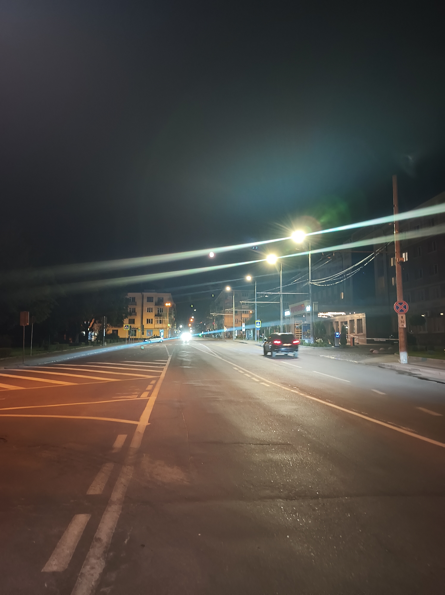 The image captures a night scene on a street in Ternopil, Ukraine. The perspective is from a vehicle looking forward, indicating a moving camera. The road is paved and illuminated by streetlights, with yellow and white lane markings visible. On the left side of the road, there are pedestrian crossings and trees. Further down the road, buildings are visible, some with illuminated windows. Two cars are present on the road; one is approaching the viewer, its headlights creating a bright glare, and another is moving away from the viewer, its taillights illuminated. The sky is dark, characteristic of nighttime, with some light pollution and lens flares from the streetlights. There are no people clearly visible in the image.