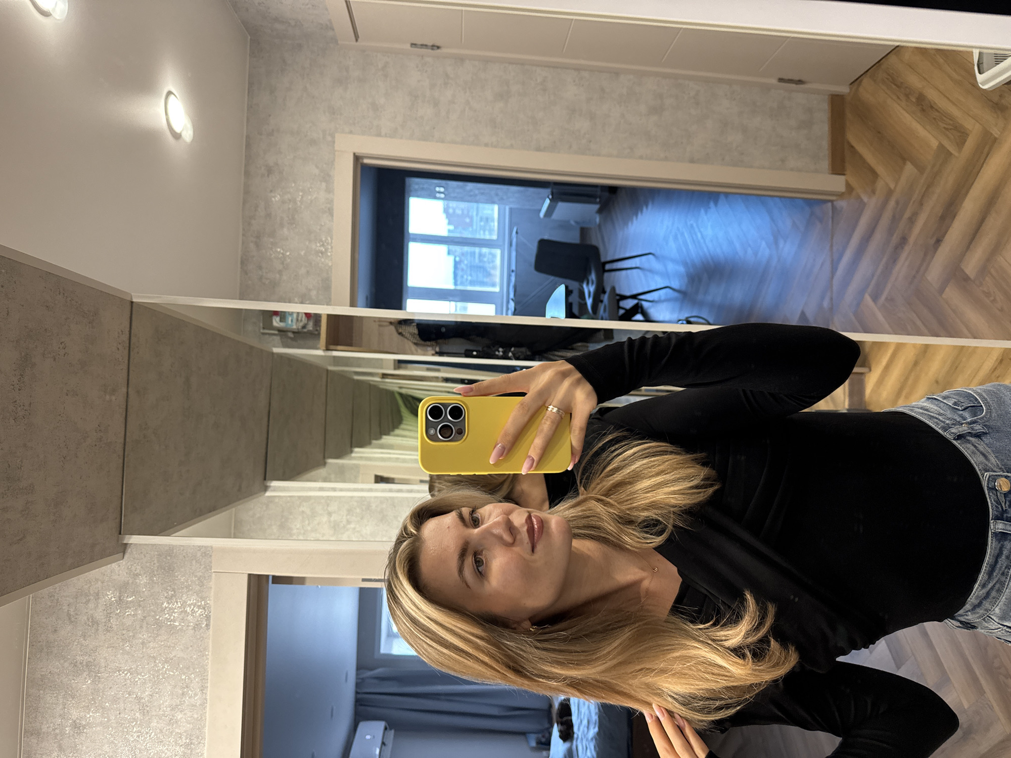 A woman is taking a mirror selfie indoors. She is holding a yellow smartphone in her right hand. She is wearing a black long-sleeved shirt and blue jeans. Her hair is long and blonde. The background reveals a hallway with light grey walls and a herringbone pattern wood floor. A doorway is visible, leading to another room with a window and furniture. The location is Unknown, Unknown.